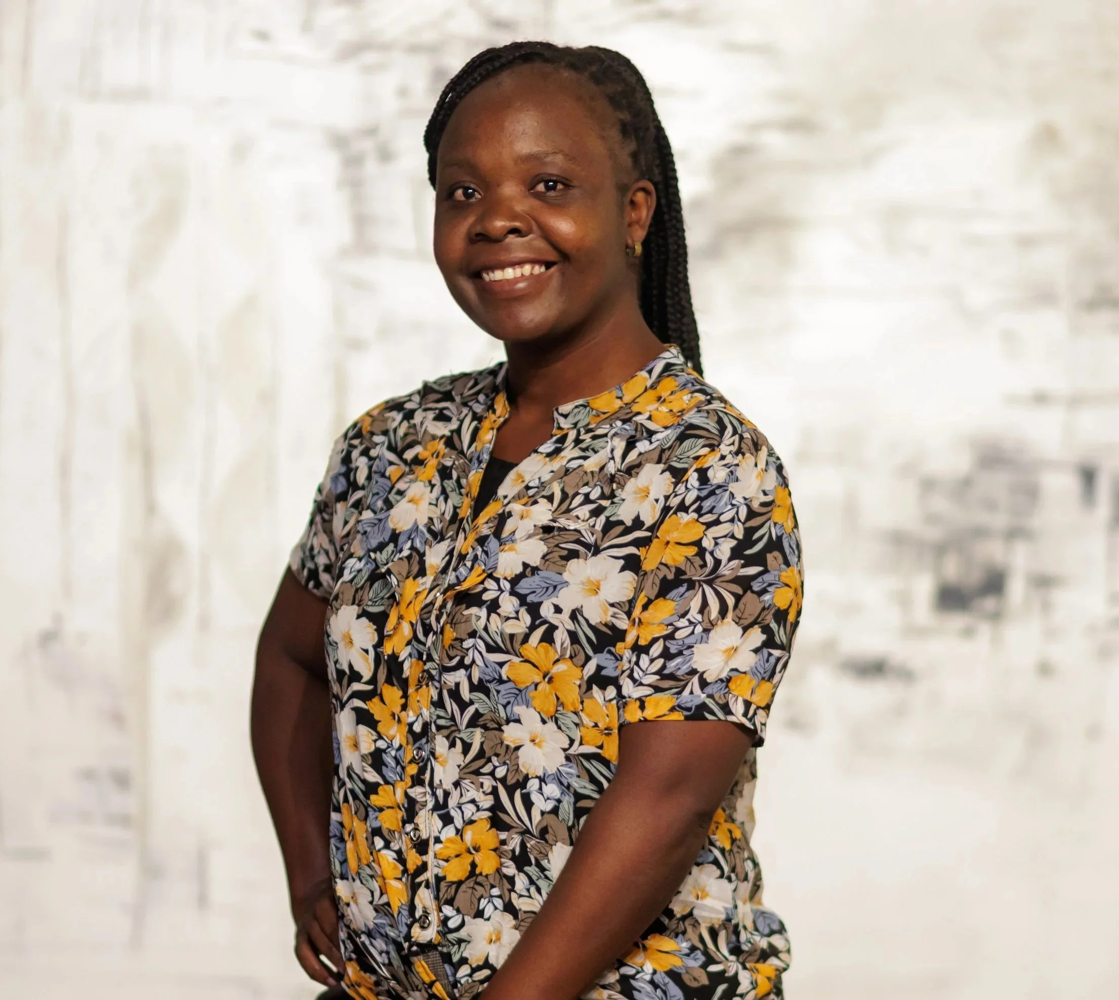 A smiling woman with dark braided hair pulled back, wearing a floral short-sleeved blouse with yellow, blue, and white patterns.
