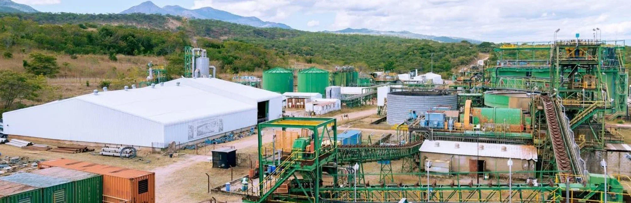A wide, elevated view of an industrial processing plant located in a hilly, rural region. The facility features a large white warehouse, green storage tanks, and various metal structures connected by conveyor belts and pipelines.