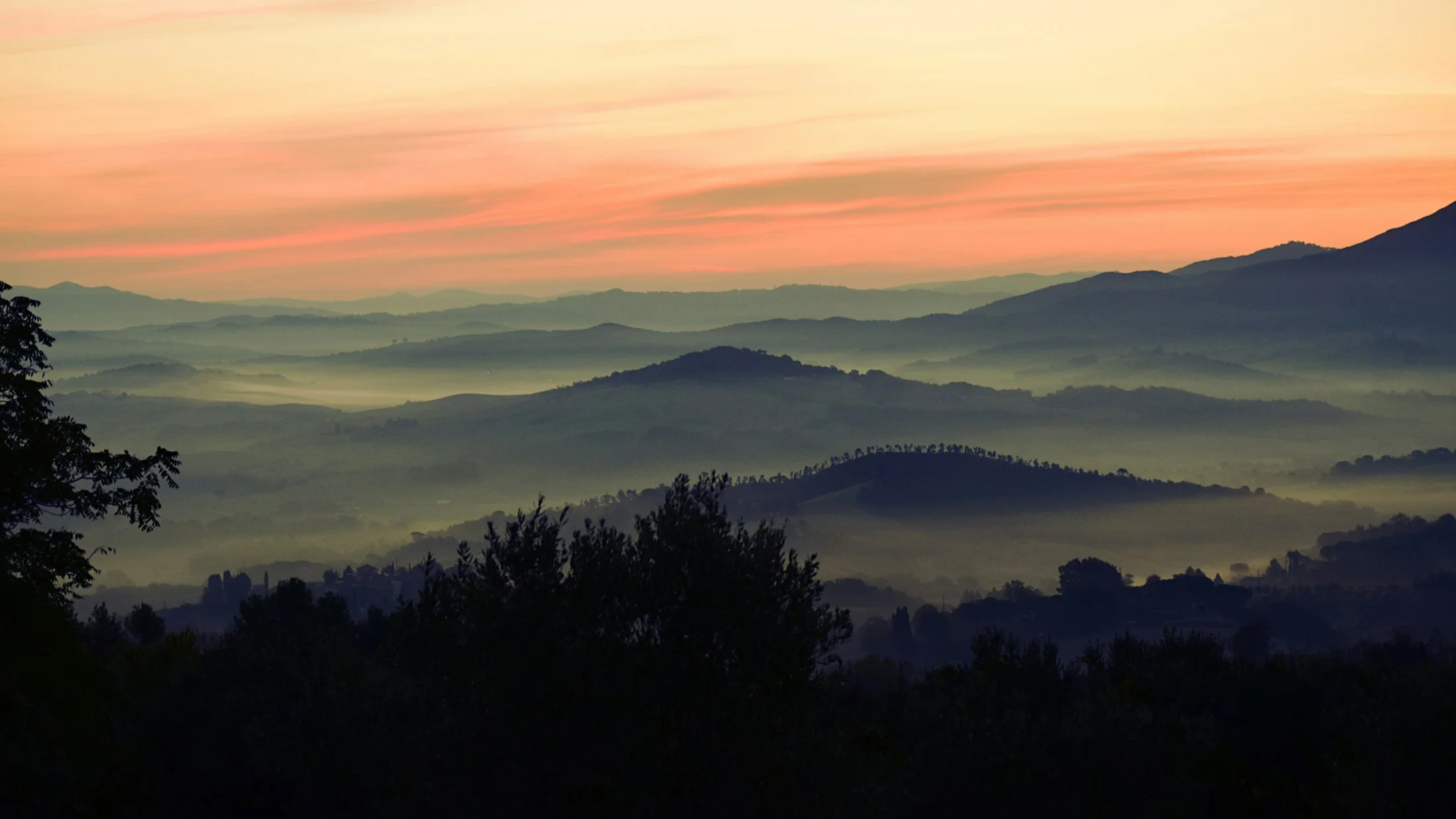 A Tuscan landscape view of rolling hills and mountains during dawn or dusk, with soft pink and orange hues in the sky and mist enveloping the distant hills.