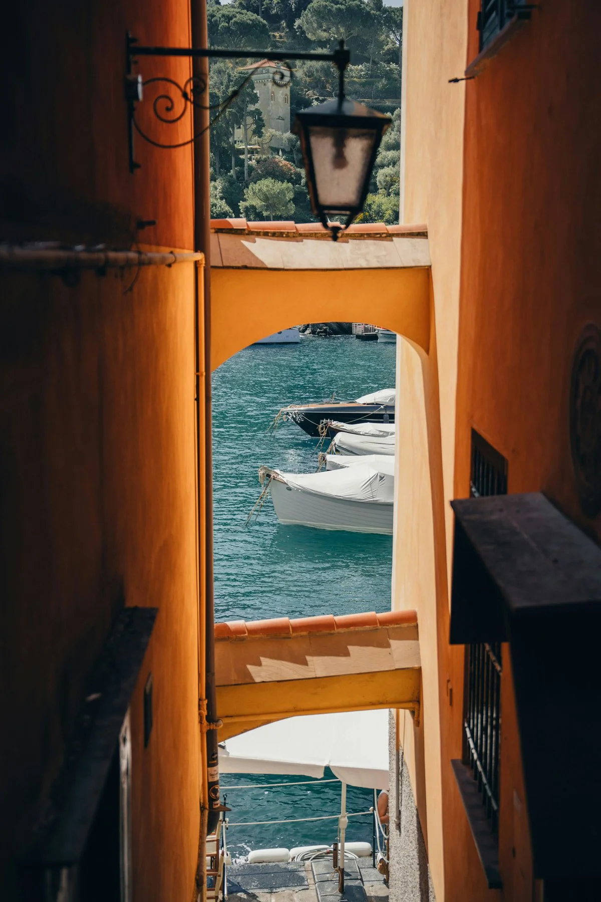 View of a Tuscan marina through a narrow passageway with orange walls with white boats on turquoise water, with a scenic hillside and a building in the background.
