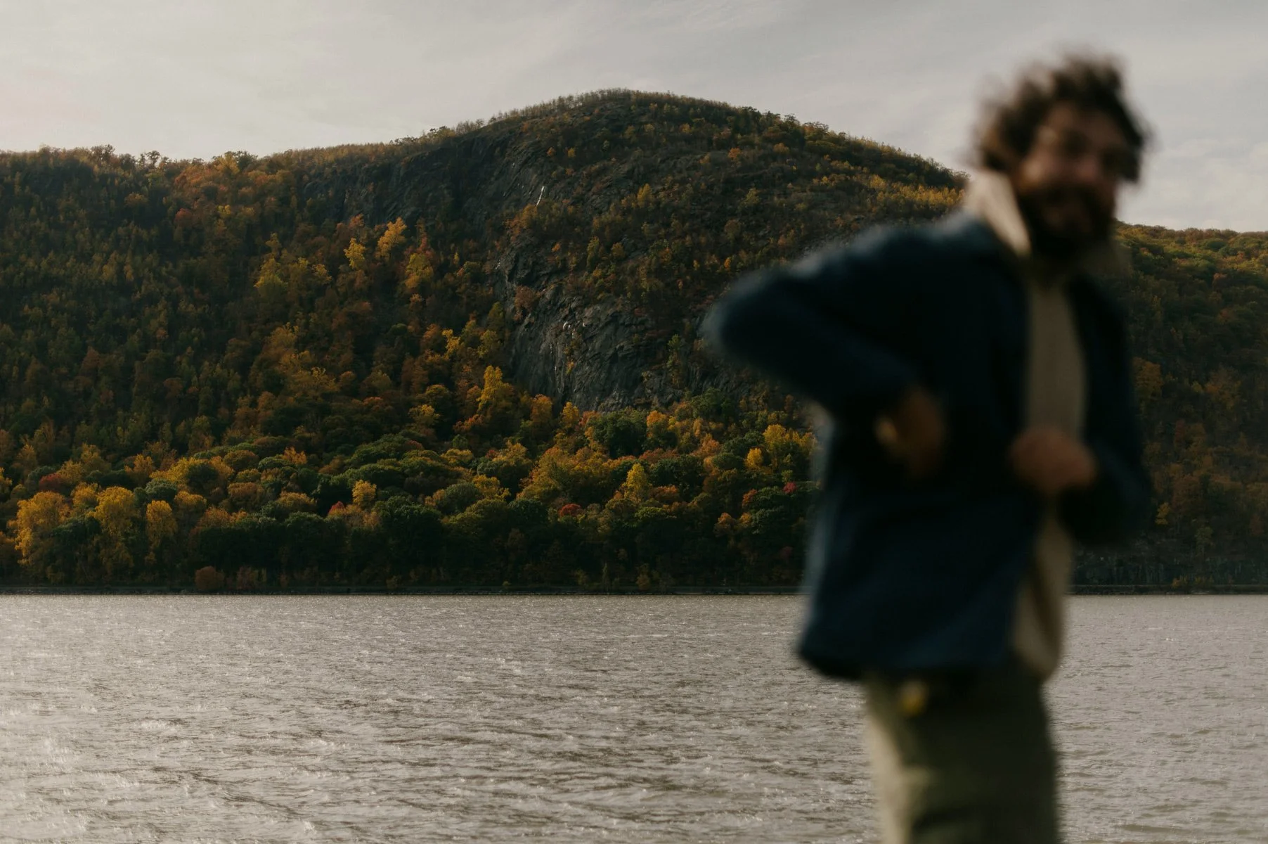 A man with curly hair and a beard standing near a body of water in Upstate New York with a forested hillside and mountain in the background during autumn.