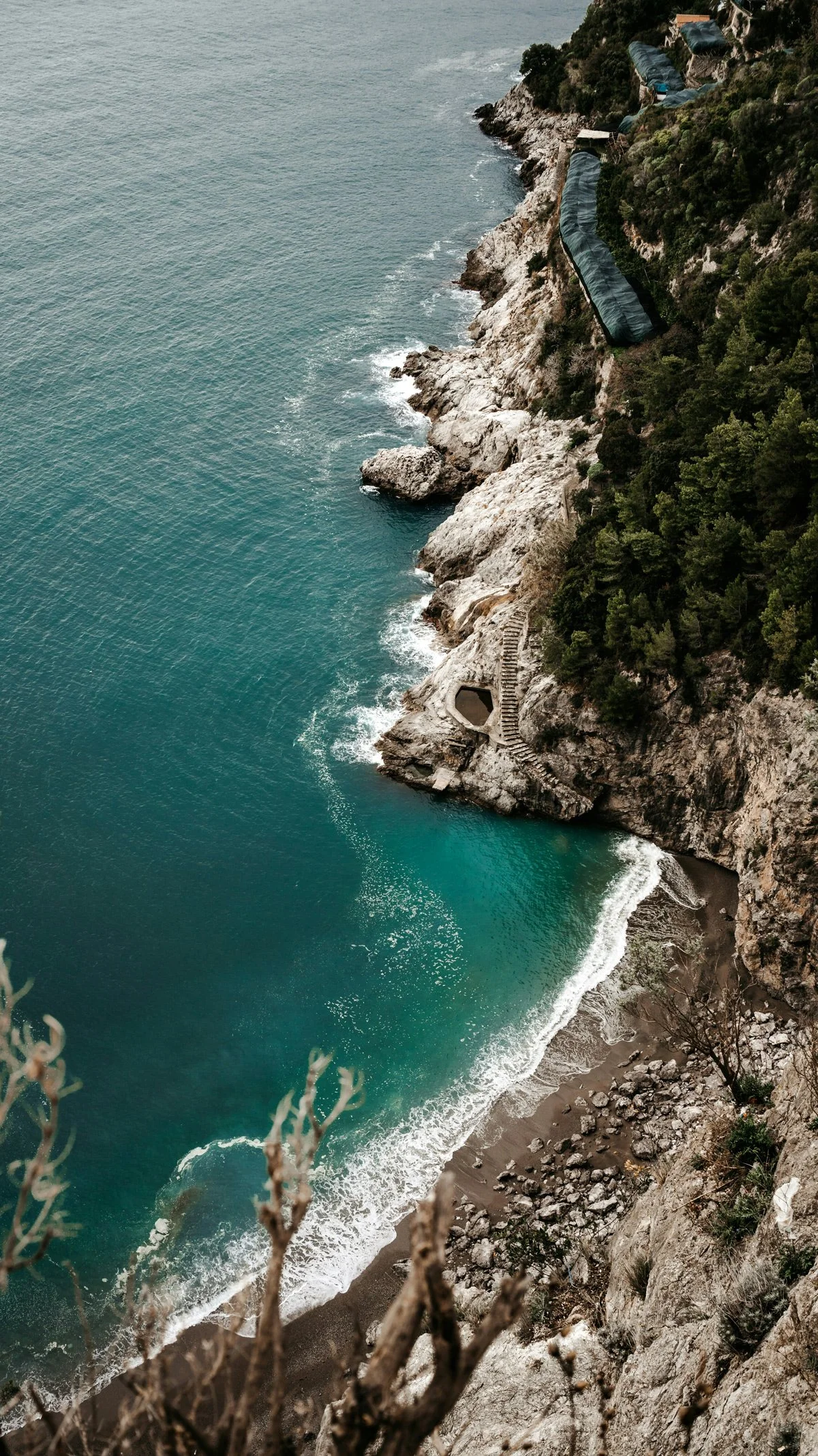 Cliffside view of the Mediterranean Sea with stairs leading down to a beach, some structures covered with green tarps on the hillside, and trees along the edge of the cliff.