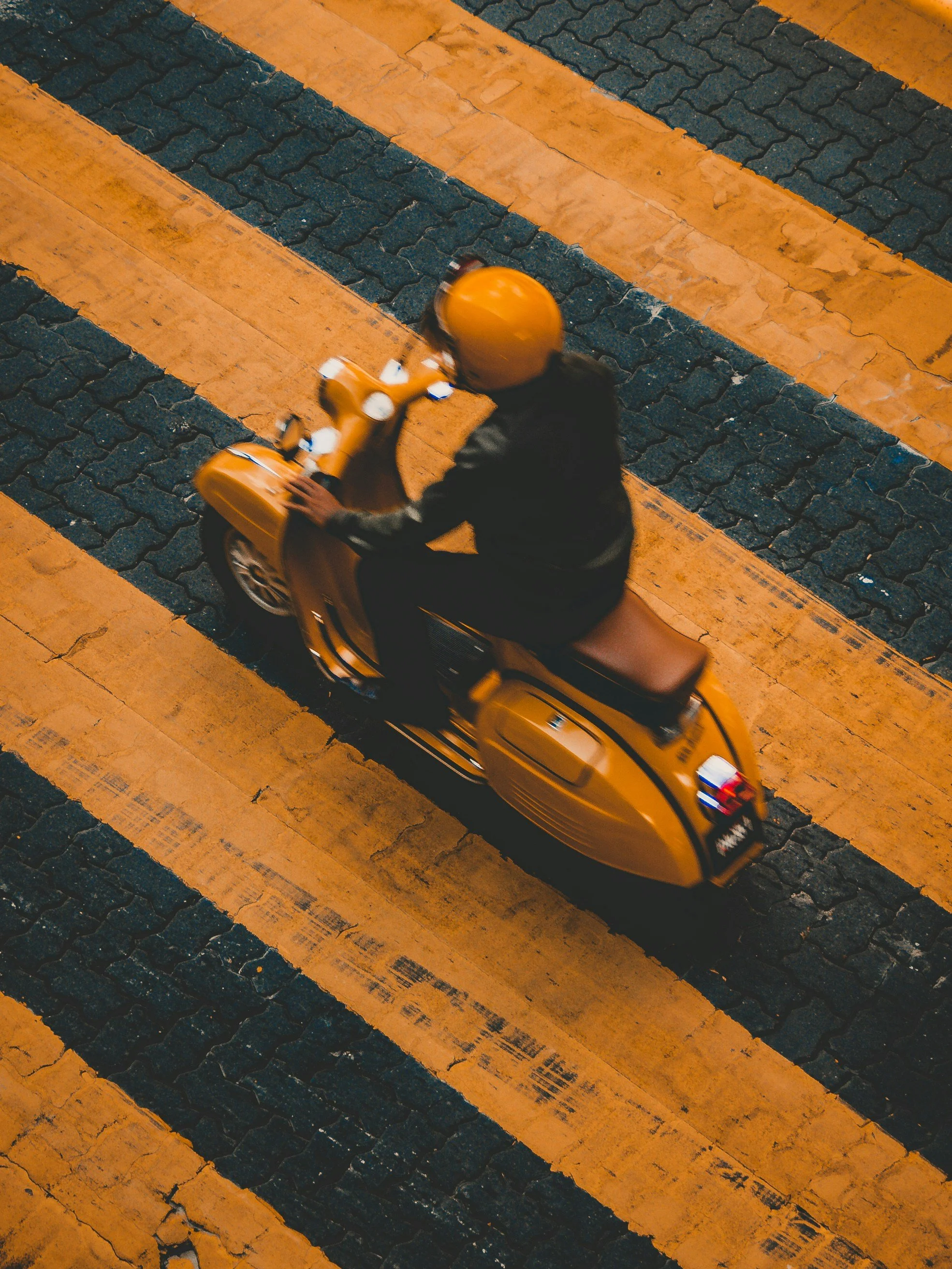 A person riding a yellow scooter across a street in Italy with yellow and black painted crosswalk lines.
