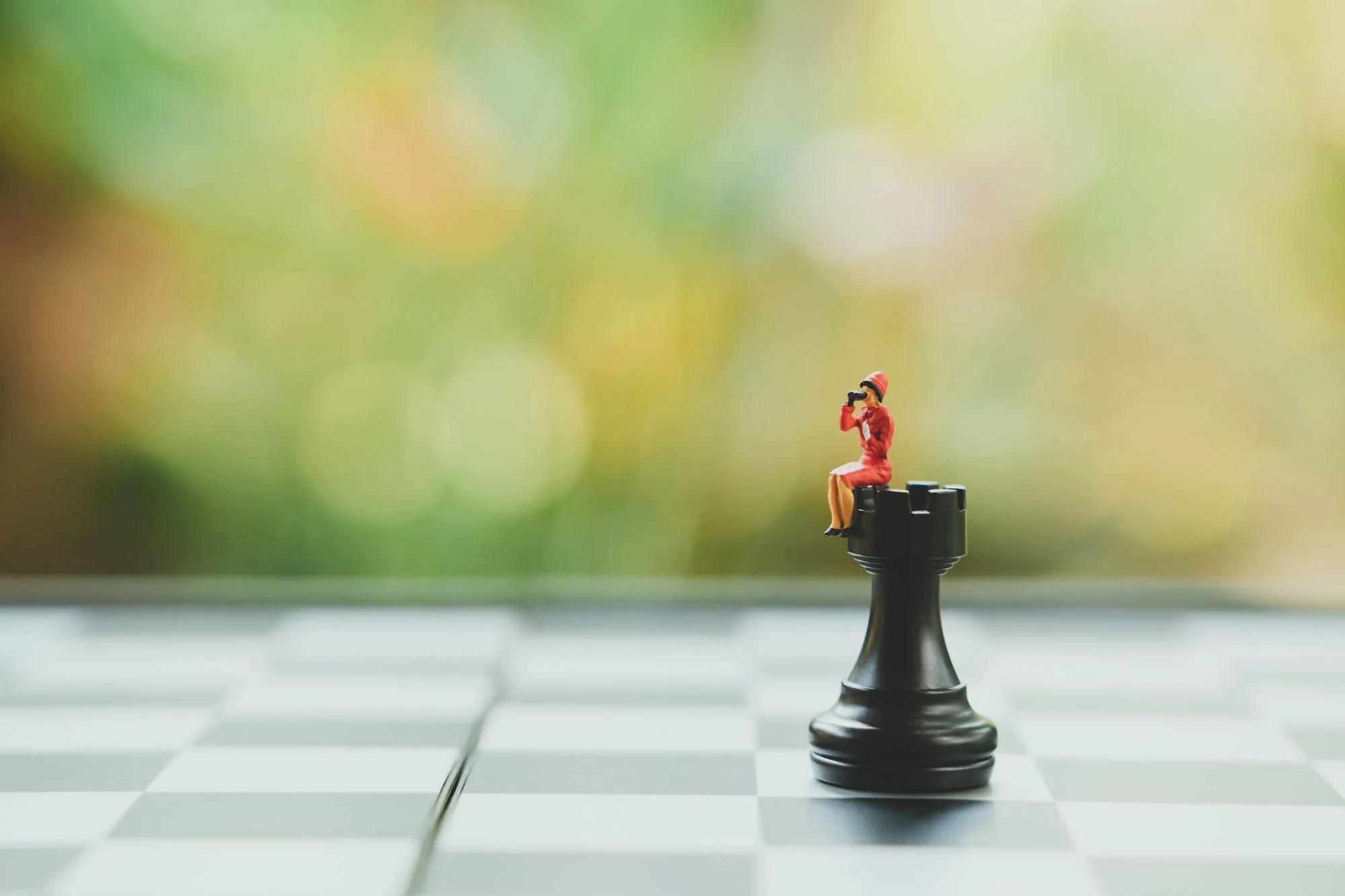 Miniature woman sitting atop a chess rook, looking through binoculars — a metaphor for ambition, strategic positioning, and the long view in executive career development.