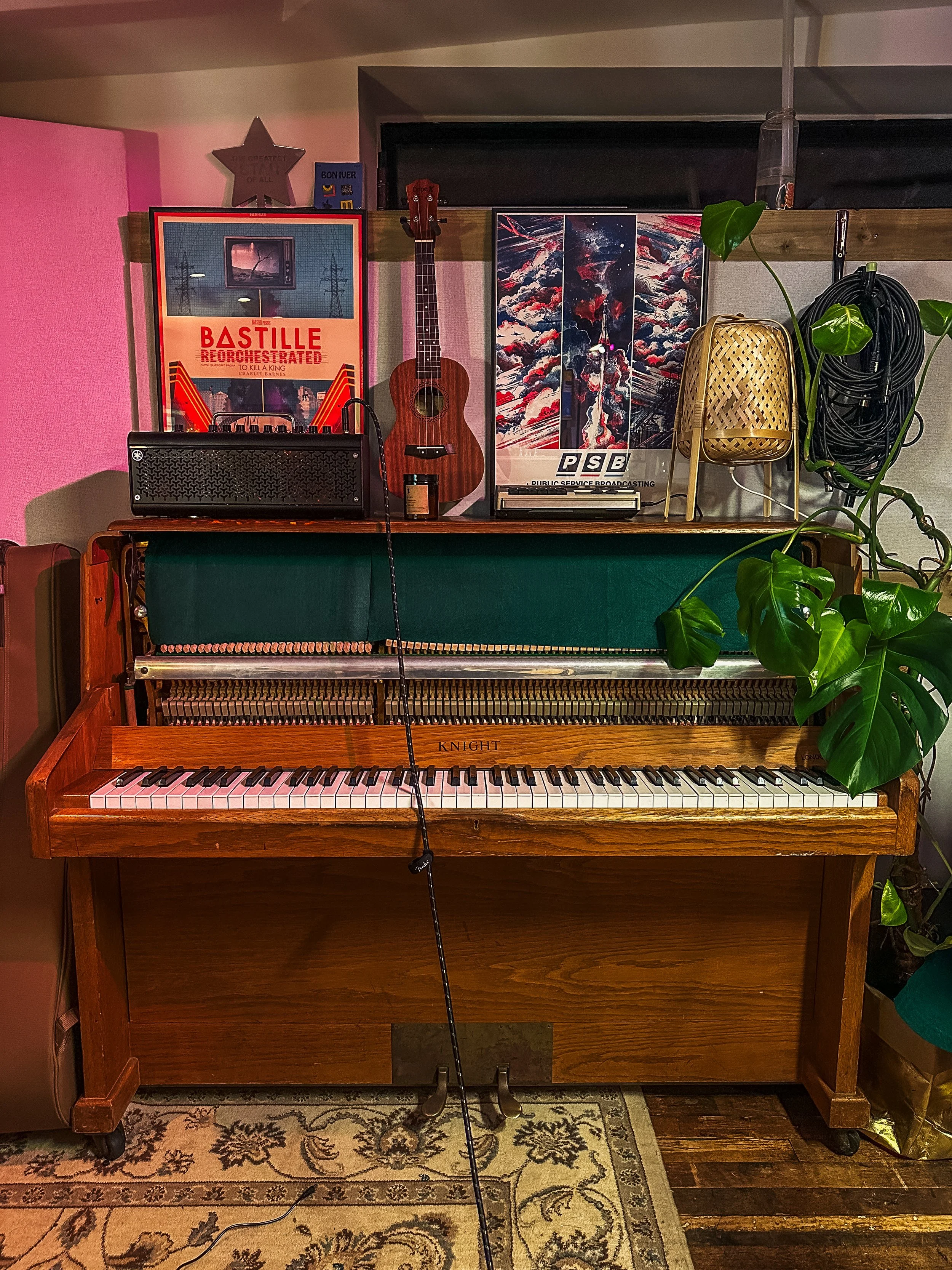 A vintage upright piano with an open top revealing the interior strings, placed on a floral rug. An acoustic guitar, a small black amplifier, and a microphone on a stand are on top of the piano. Behind the piano, there are posters, plants, and a roll of black cable.