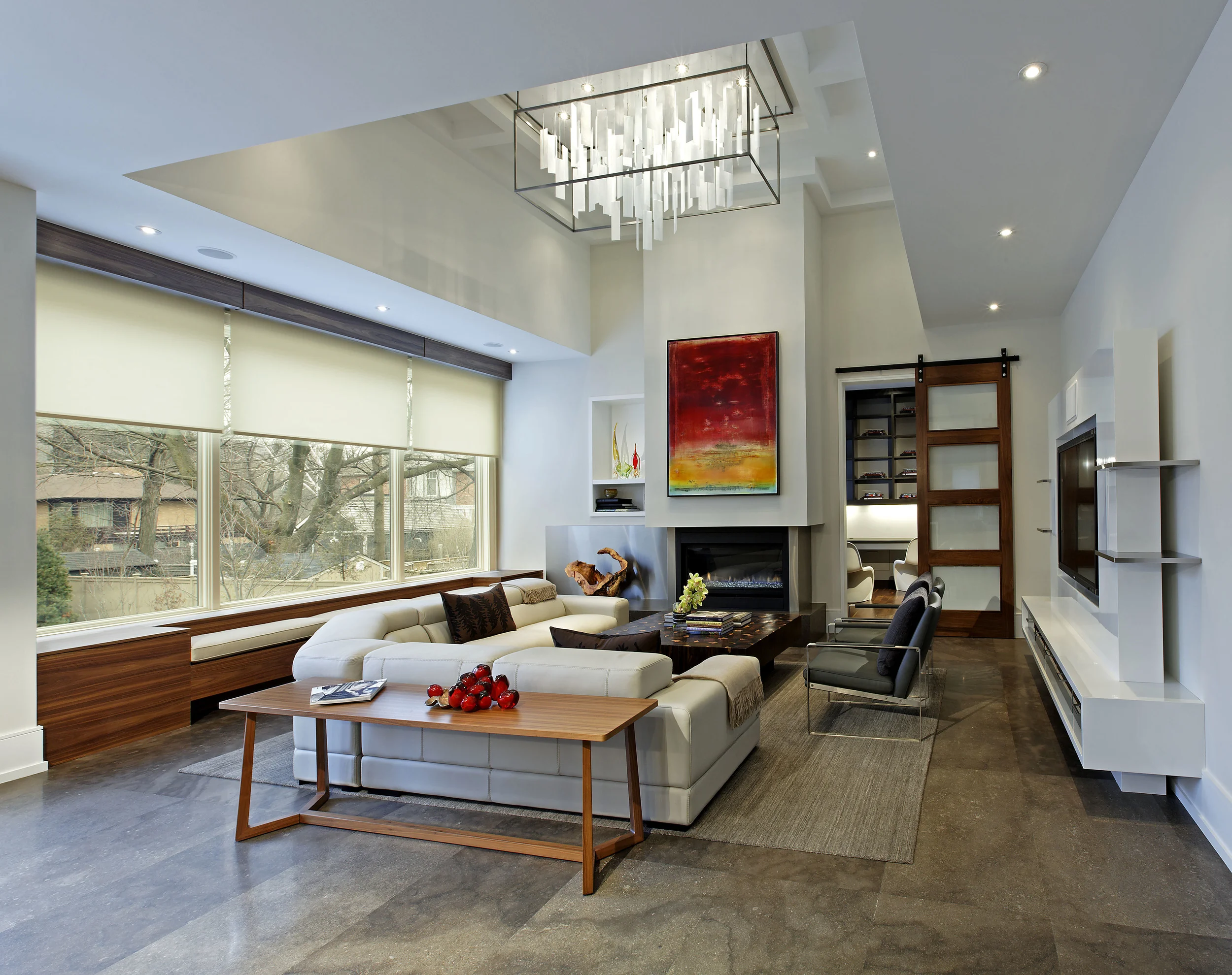 This room epitomizes the inviting, cozy, family-oriented environment created within a pared down, modern context. Its layering and sculpting includes seating and storage units tucked under the windows, below carefully planned bulkheads and a coffered ceiling and custom light fixture.