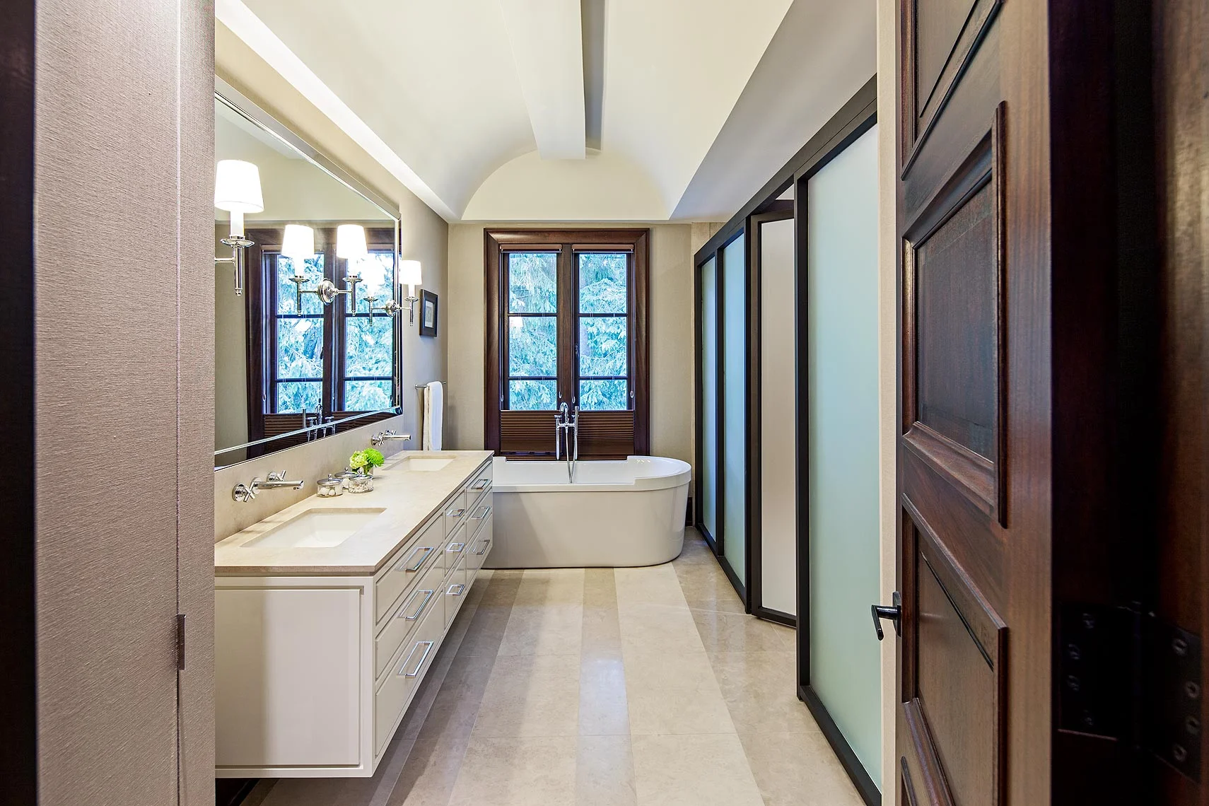 The barrel vaulted ceiling with recessed lighting emphasizes the serene and orderly nature of this peaceful retreat. Bronze-framed frosted glass partitions conceal the shower and toilet room. Top down/bottom up, bronze-toned fabric blinds create privacy while preserving the view of the lush landscape surrounding this river valley home. Grasscloth covering the foreground storage cabinet adds an unexpected textural element.