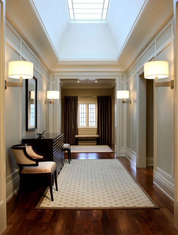 This client’s North American achievement and European heritage were celebrated in a legacy home in which each room has its own mood, united by a common richness. The long upper hall’s rhythmic pattern of doorways, arches and sconces is bathed in light from one custom polished nickel high lens. To achieve symmetry, the three windows were draped to look like two.