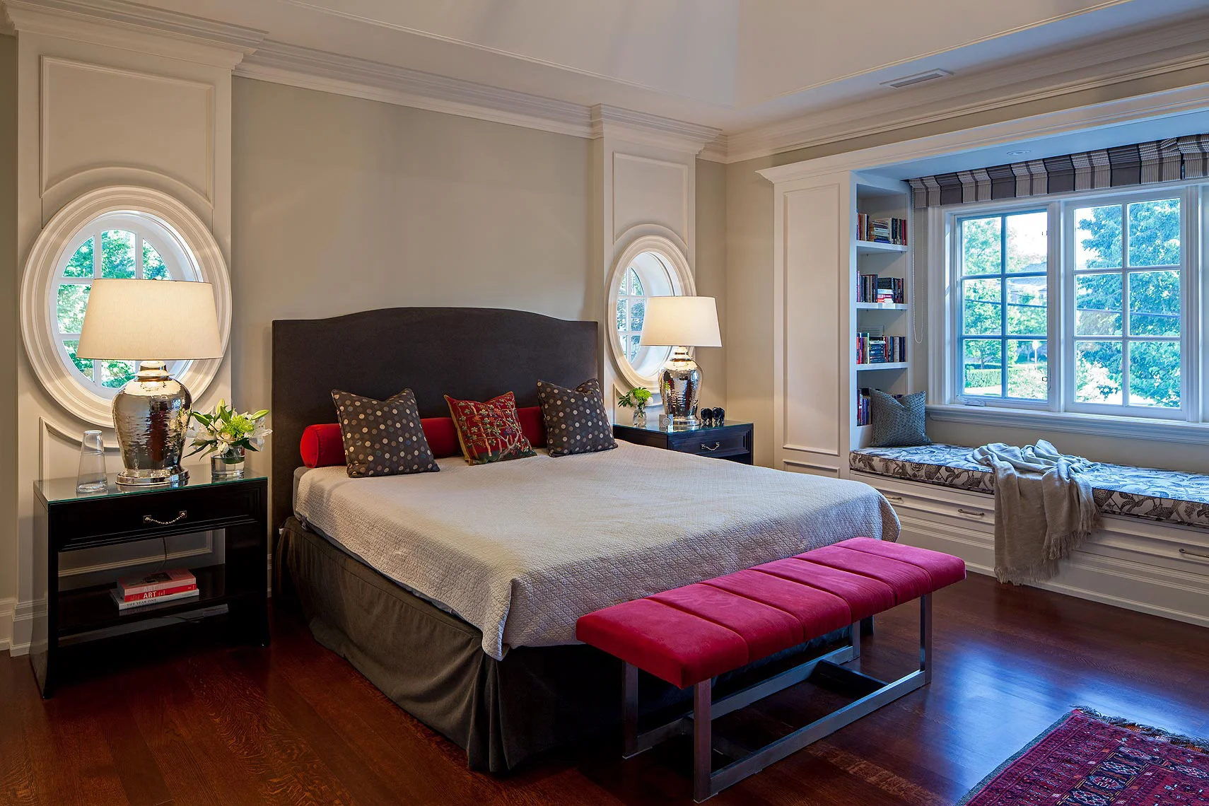The upholstered headboard is perfectly framed by the panelled wall, which incorporates two oval windows above extra-wide cabinets. The window casings were designed to conceal pull down blinds for privacy. The adjacent wall was built out to incorporate storage and an 11 foot long daybed under the huge window. The serene colour scheme is accented with red elements such as the bench cushion and the throw pillows’ large scale prints, which reference the antique fabrics and carpets found elsewhere in the home. Nickel finishes on the lamps and hardware add sparkle.