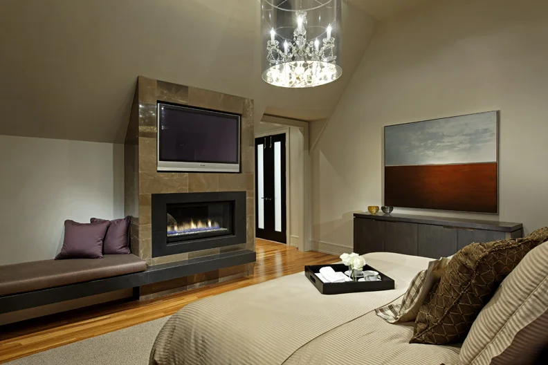 Deep tones add intimacy and richness to this bedroom, in which the fireplace, TV and fabric-wrapped bench form one functional, cohesive unit tucked below ceiling slope.