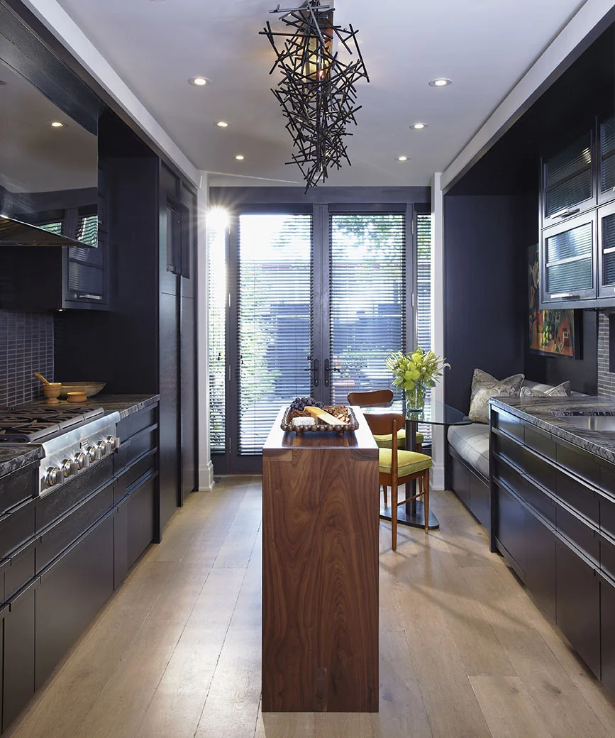 Working with a long and narrow space, a galley-style kitchen was created with integrated, hidden appliances. A 16” wide custom designed island is the perfect plating size for 6 dinner guests. Upper cabinets flip up and out of the way for easy access while cooking. Lower pull-out cabinets have integrated handles for a streamlined look. The richness of the charcoal colour, combined with the oak floors, creates a warm cozy feel. Clean lines are complemented with a striking custom light fixture by Zac Ridgely.