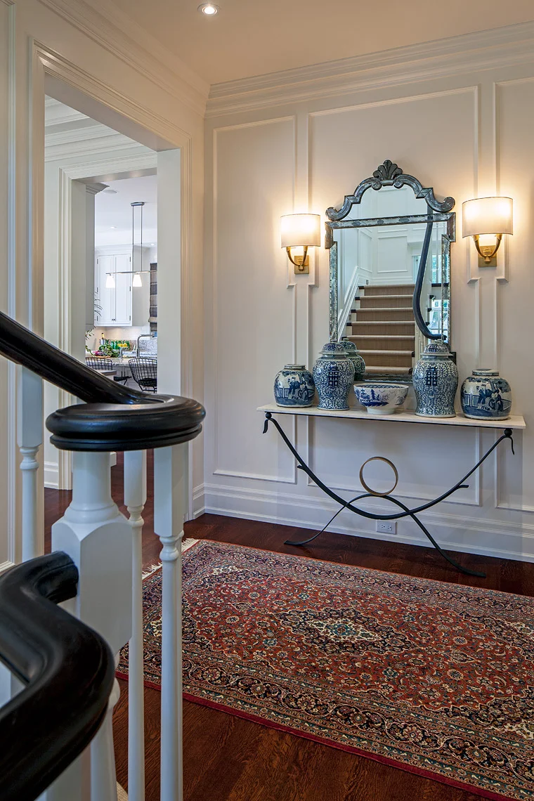 Set against the elegantly designed traditional paneling and millwork and antique carpet, the diverse collection of furnishings at the base of the stairs emphasizes that a modern family occupies this traditional home. Flanking the traditional mirror-on-mirror centrepiece, elegant sconces fit perfectly into the paneling detail. The curves of the shades and of the contemporary, custom-made, forged iron and gilt console compliment those of the sinuous stair.
