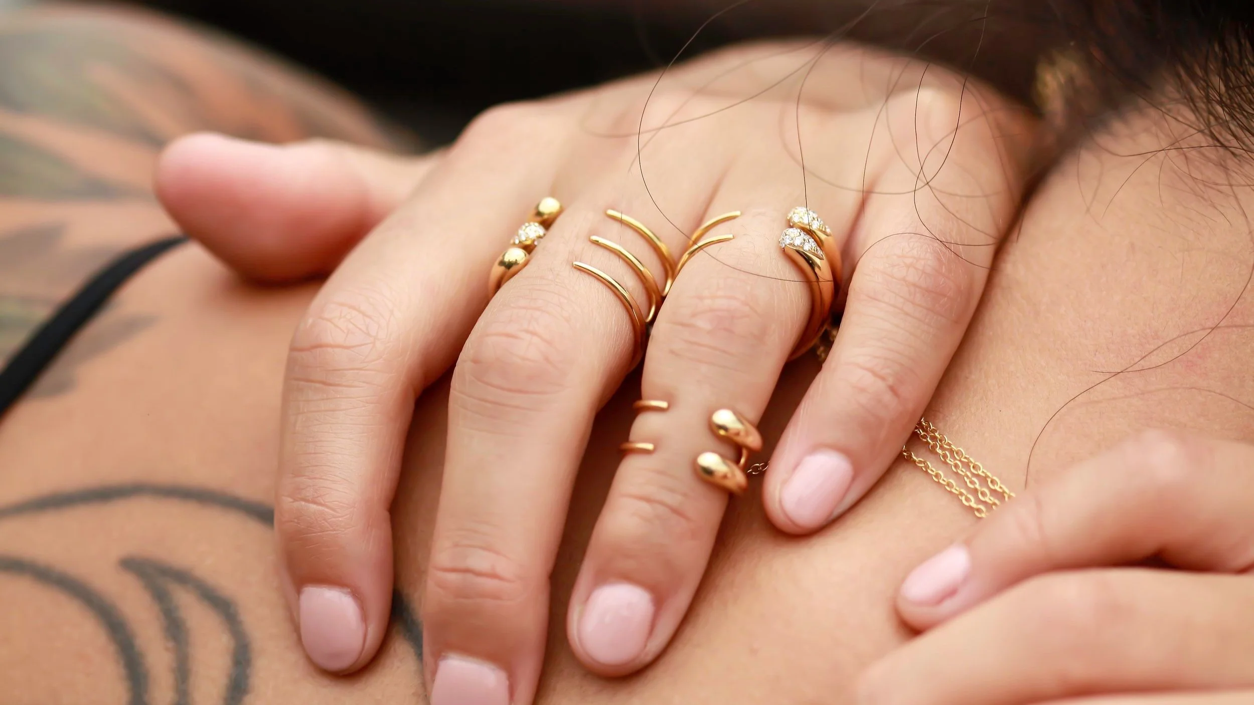 Close-up of a woman's hand with multiple gold rings, some featuring diamonds, resting on her chest near a tattoo.