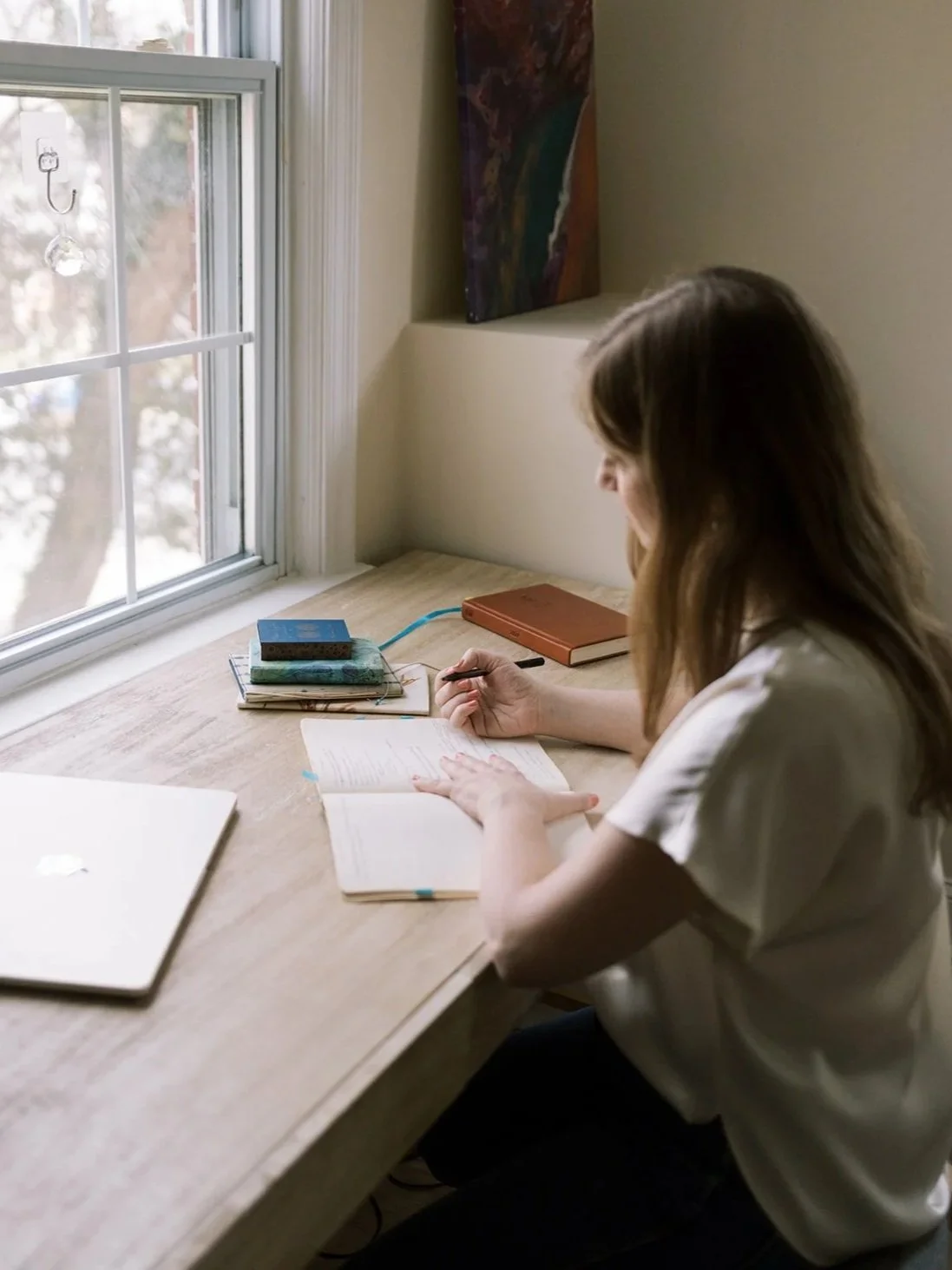 Person writing in a notebook at a wooden desk beside a sunlit window, with books and a laptop nearby