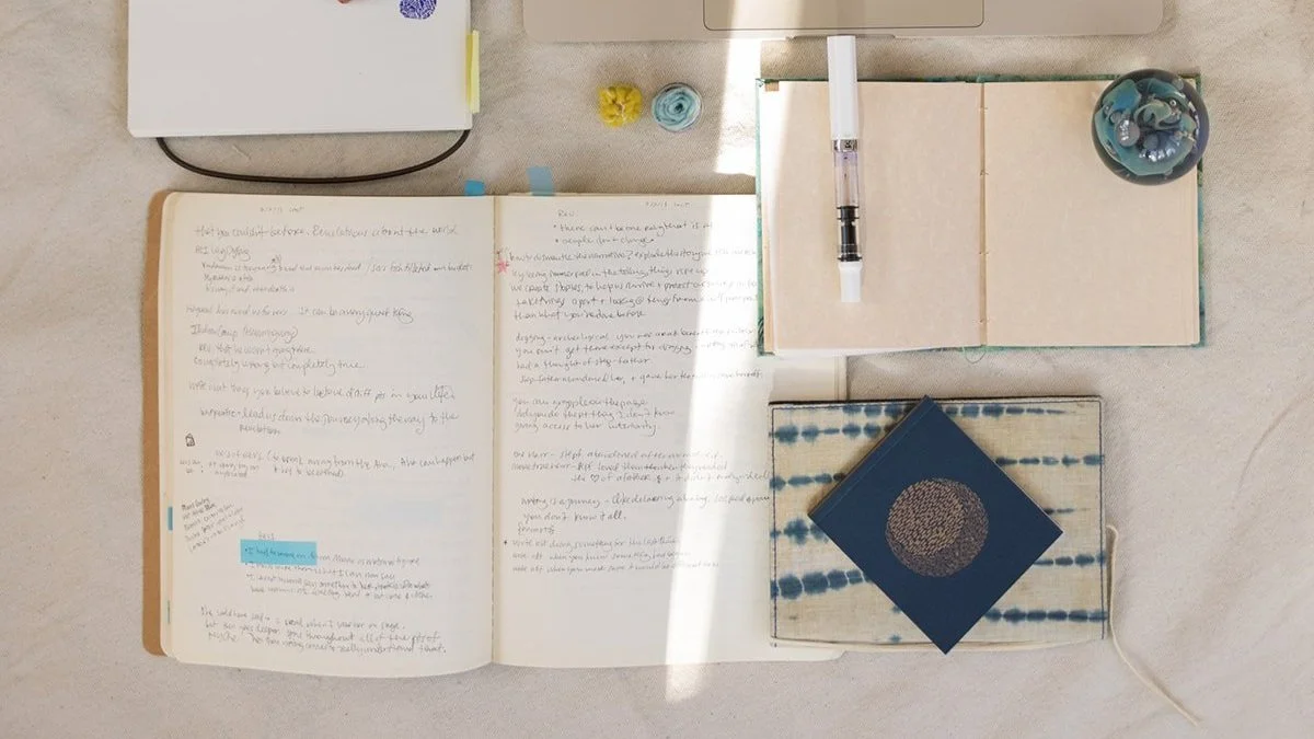 Overhead view of an open handwritten journal, sketchbook, notebooks, pen, blue geometric card, and small decorative objects on a light fabric surface