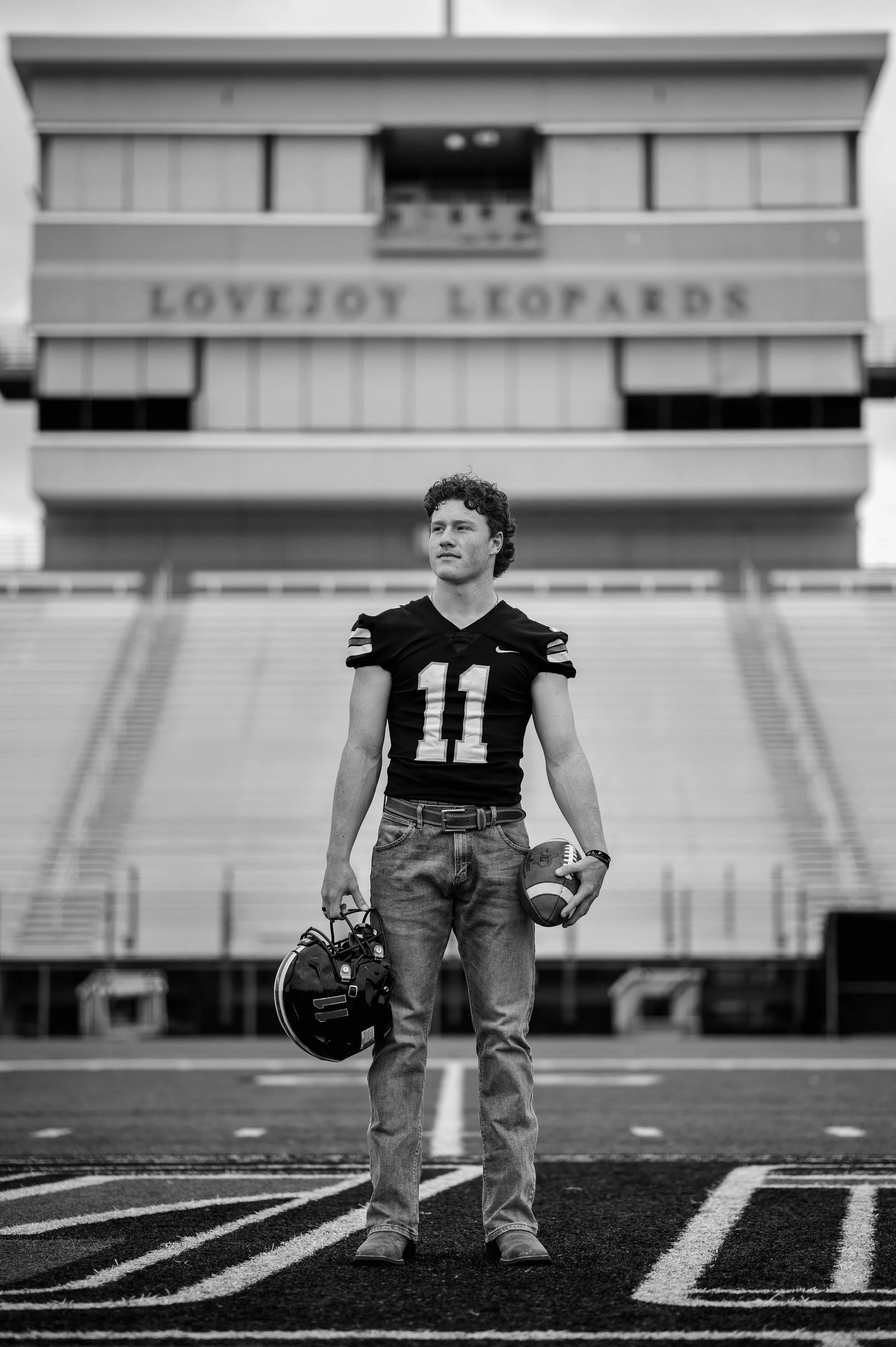 Black and white full body shot of varsity football player in front of bleachers standing on the 50 yard line, holding a football in one hand and his helmet in the other.