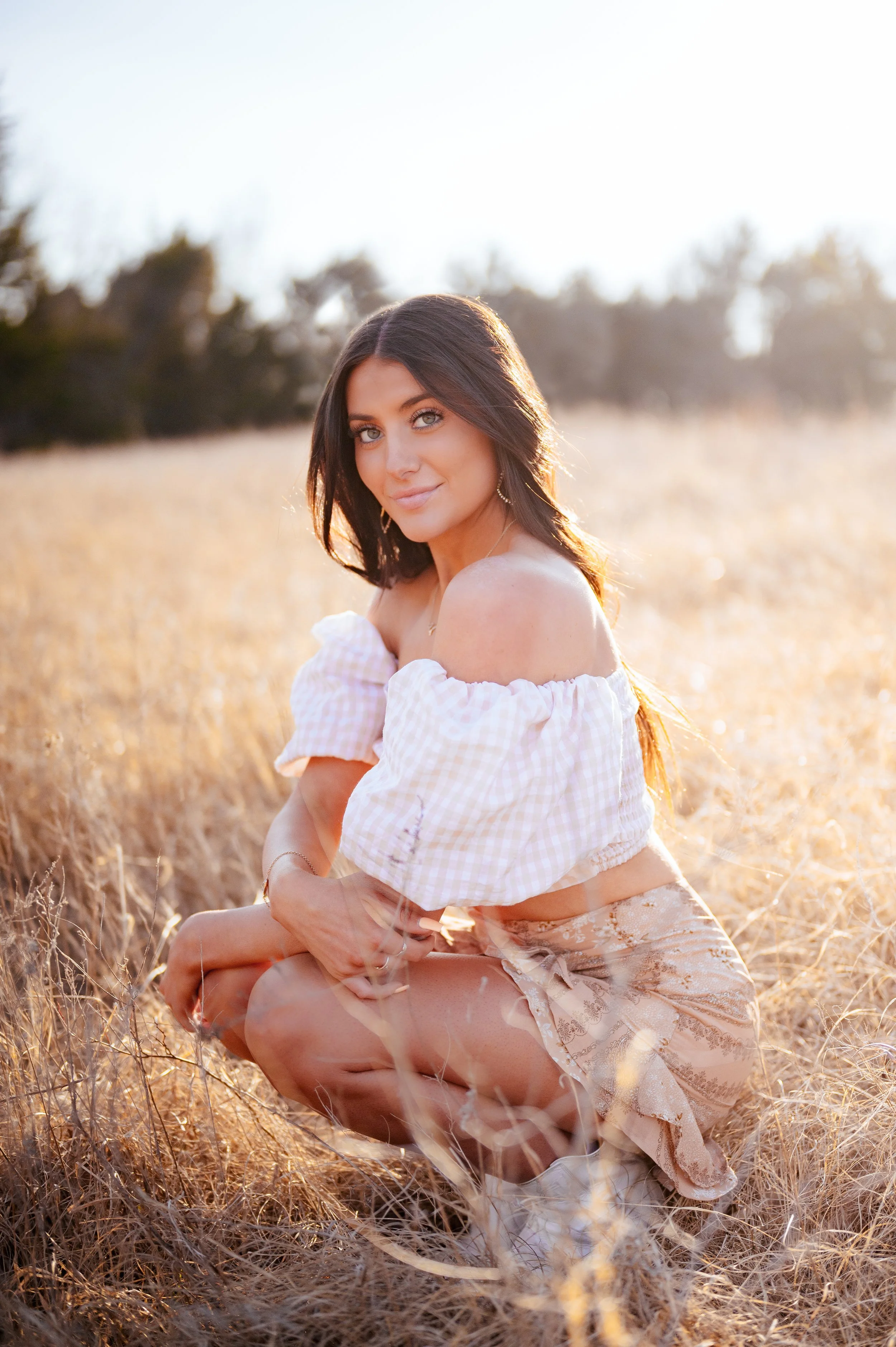 Brunette high school senior girl poses squatting in an open field with her arms crossed out in front of her over her knees. She wears a white top and tan skirt and there's a treeline in the distance.