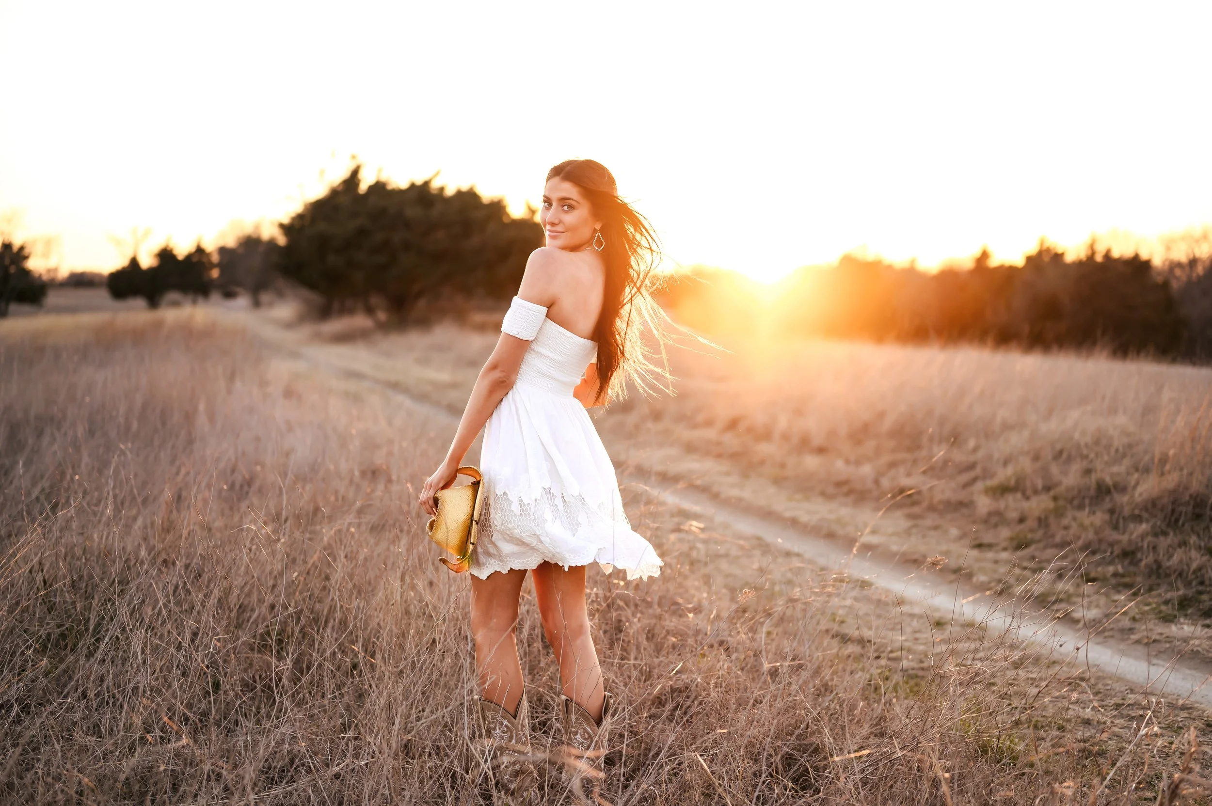 A smiling high school senior girl stands outdoors in an open field with tall grass, wearing a white sundress and holding a cowgirl hat. The background features a tree line and sunset.