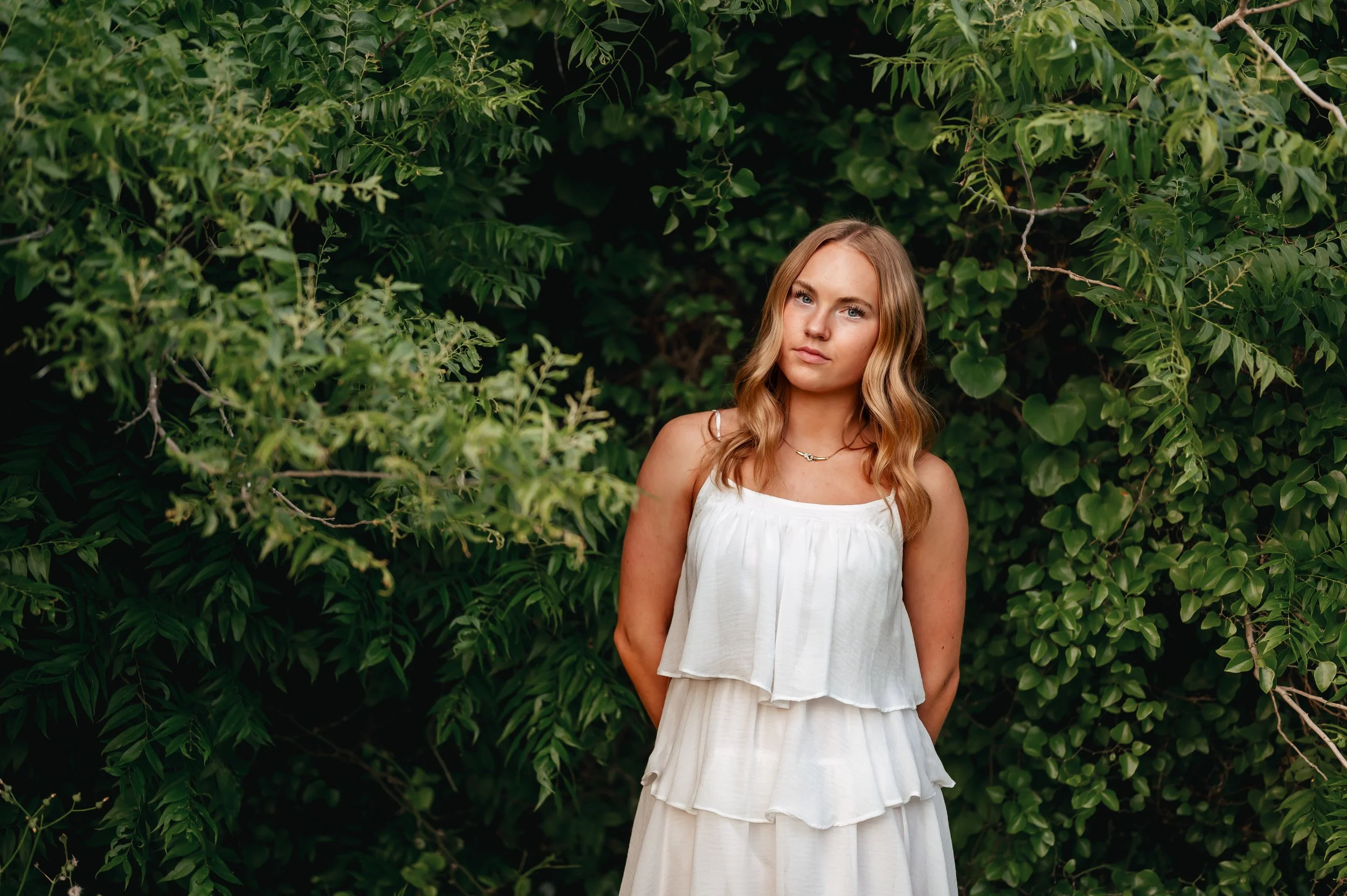High school senior girl in white sundress poses with arms behind her back and a straight face, in front of green trees.