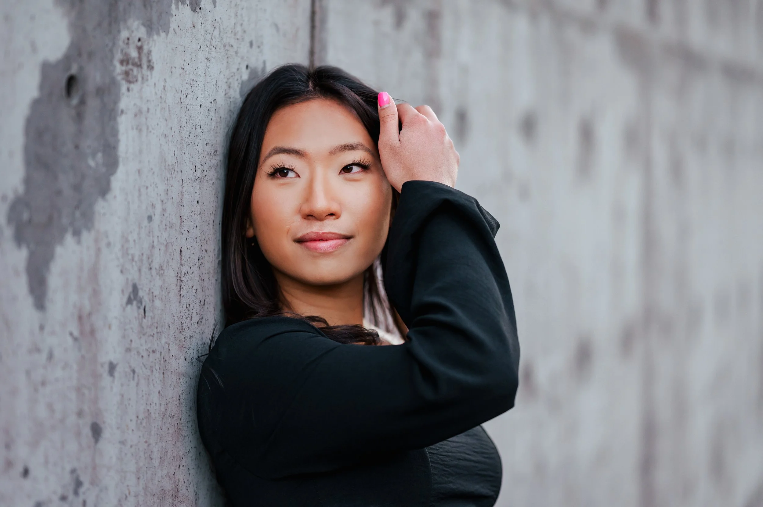 Senior girl with dark hair and black top poses in front of gray cement wall for a closeup shot, and her right arm crossing over to move her hair.
