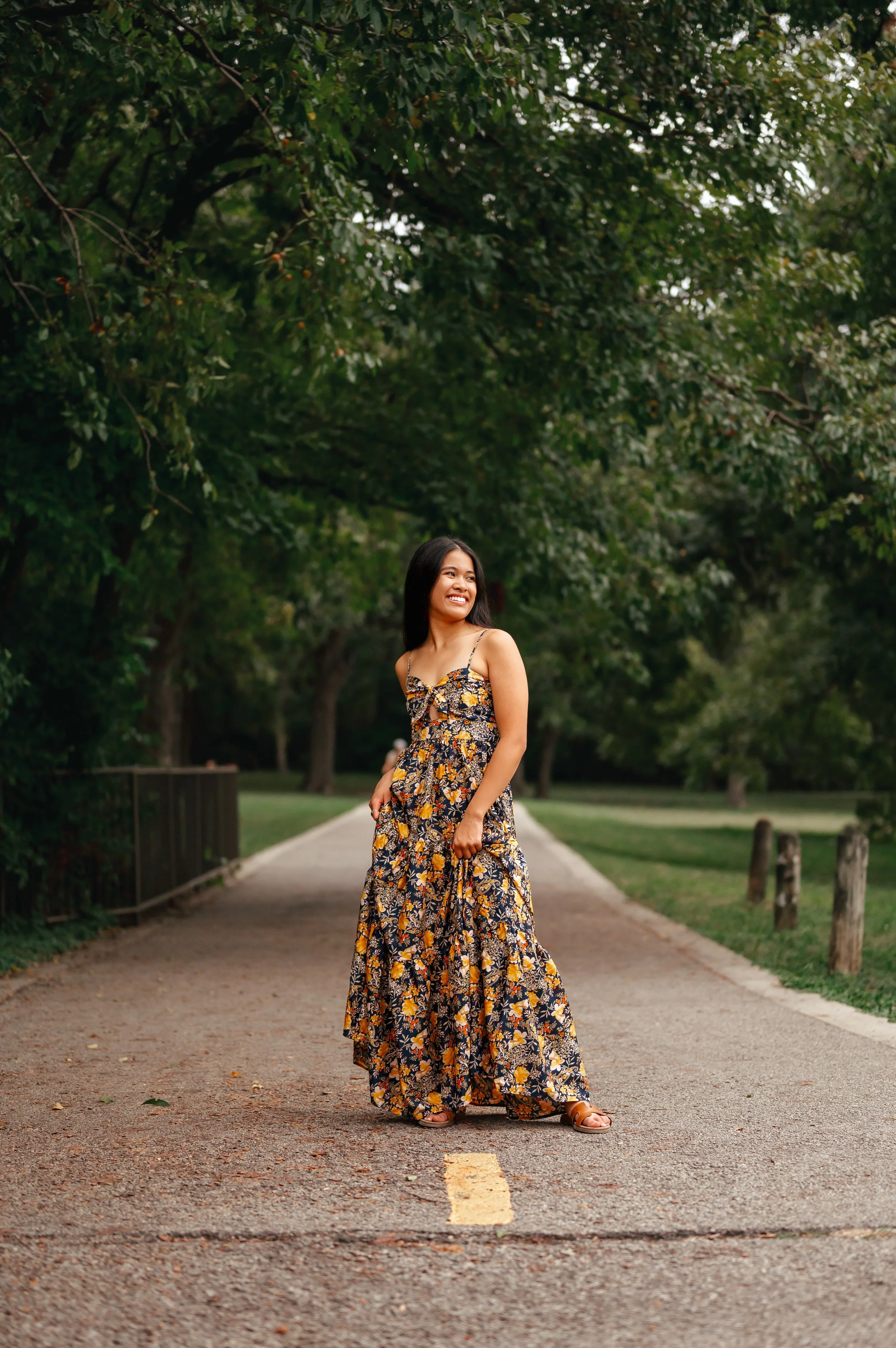 High school girl stands on road on yellow stripes, in a fully body shot featuring a long floral dress and green trees in the background.