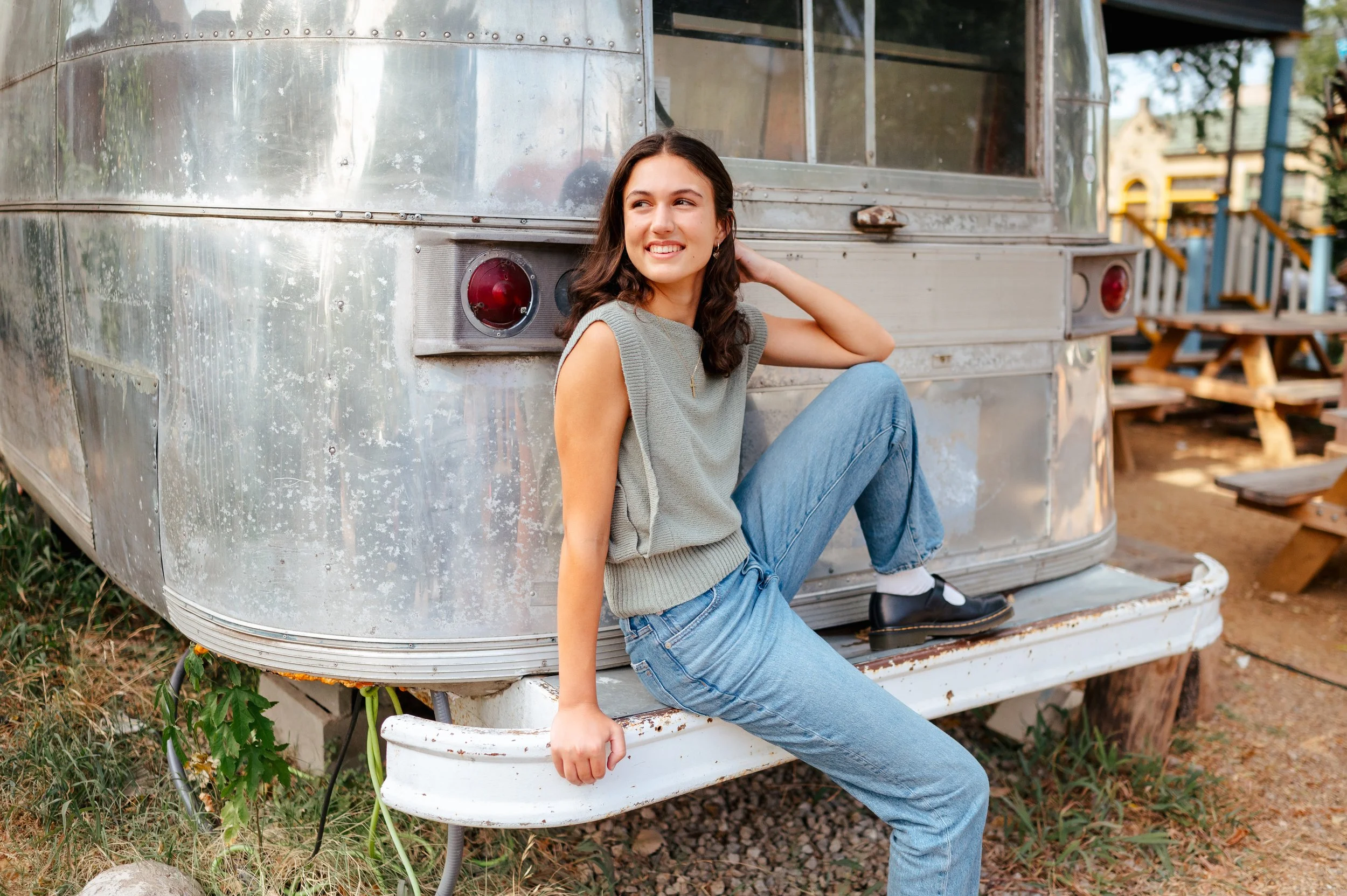 High school senior girl poses sitting on the back of an airstream trailer. She wears blue jeans and a sleeveless gray top