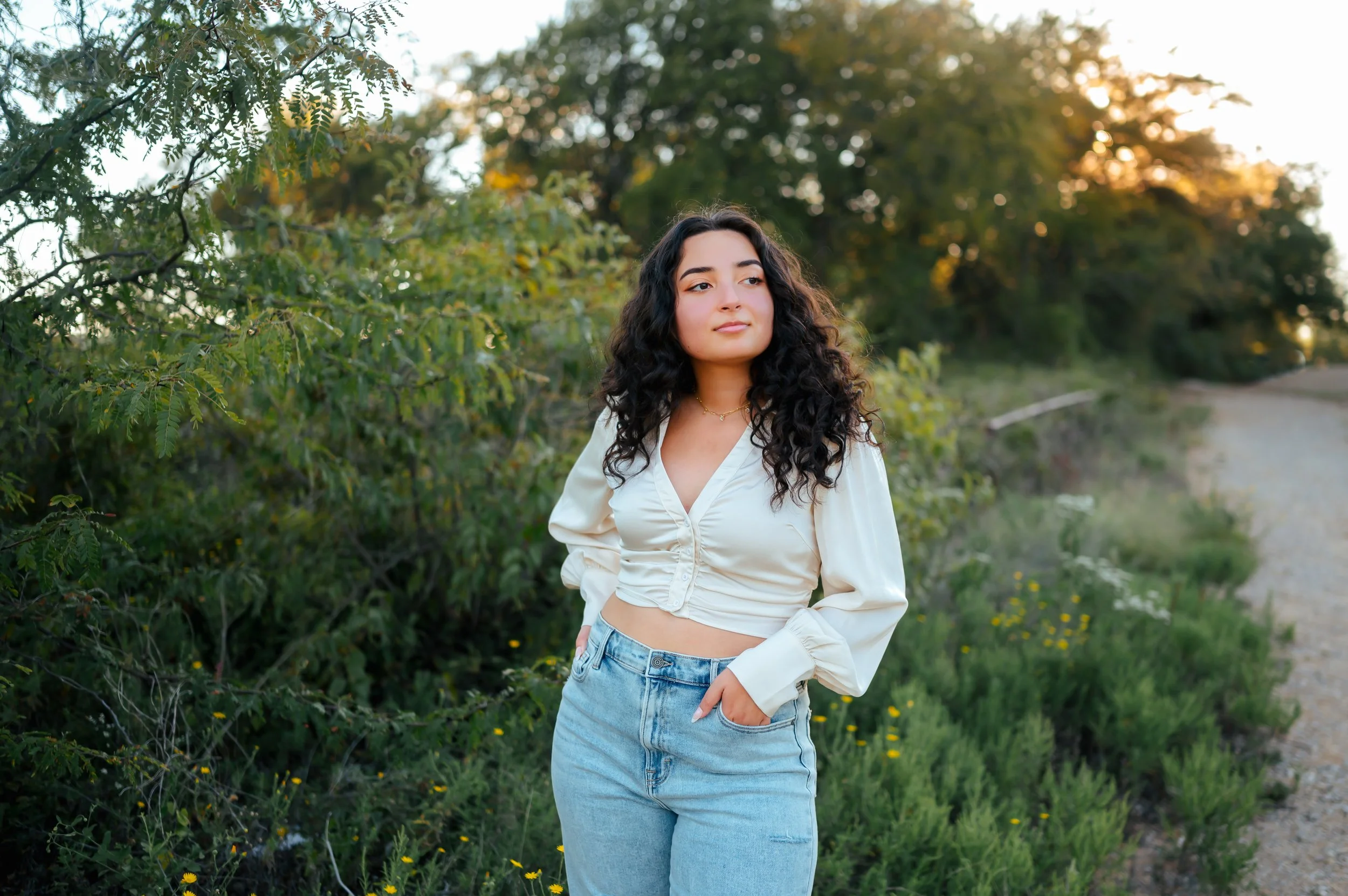Senior girl with dark curly hair stands with hands in pockets in green shrubbery, next to a gravel road with a sunset in the background.