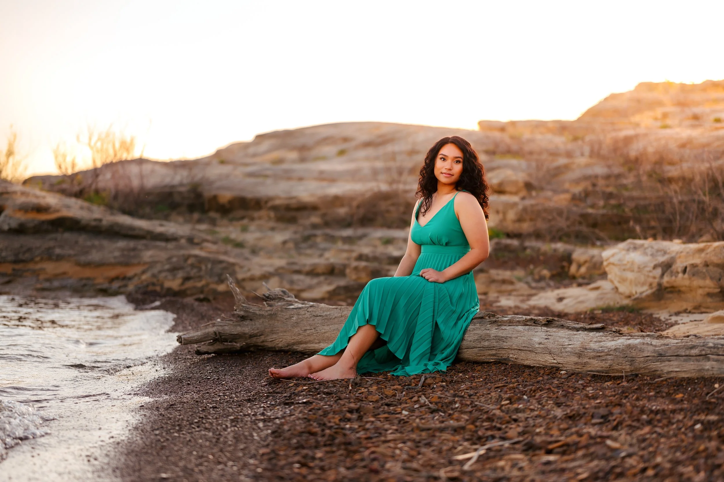 Teen girl in emerald green dress sits along rocky shoreline with sunset light in the background