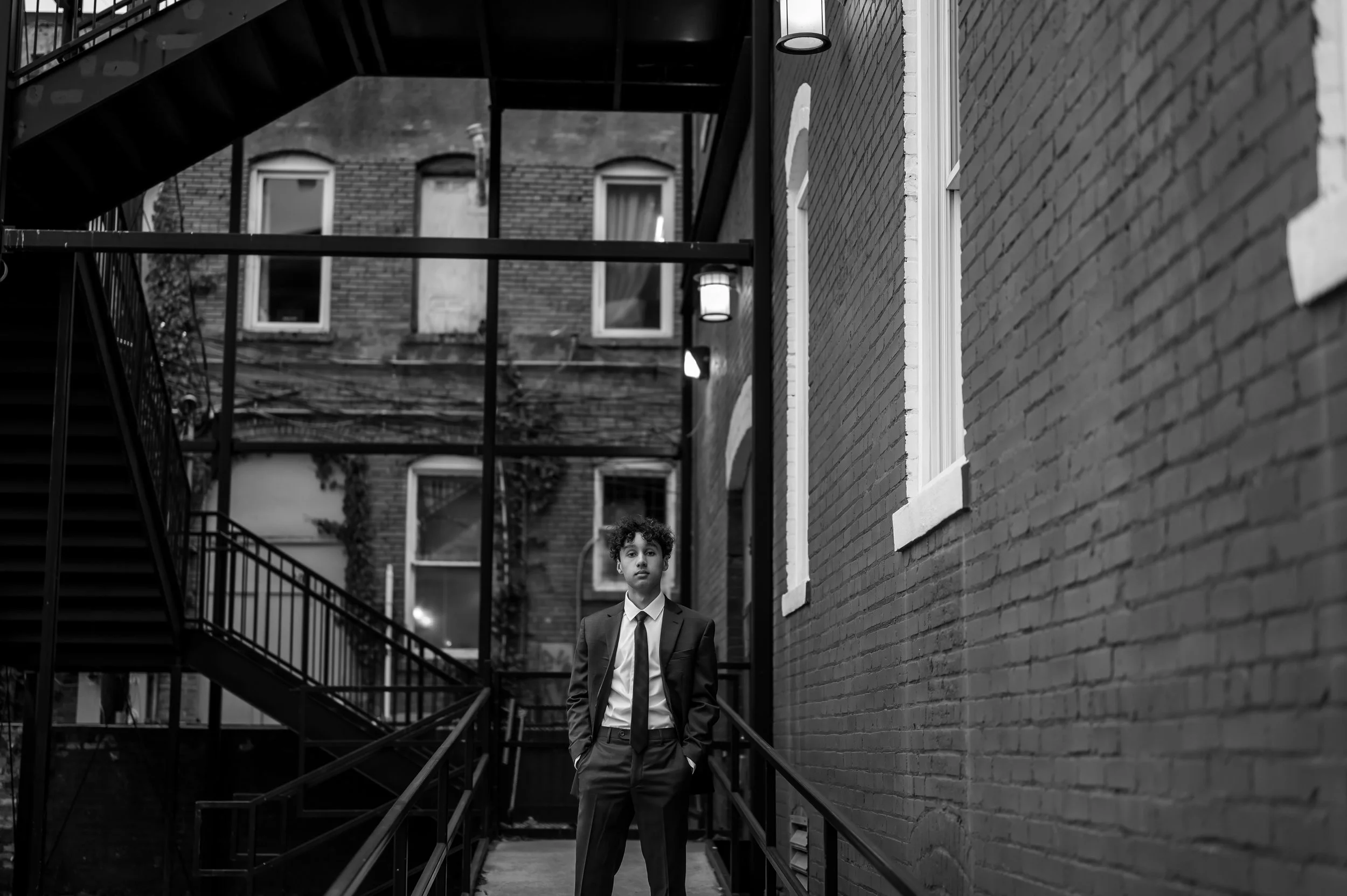 High school senior boy poses standing wearing a suit on a cement ramp with buildings beside and behind him. The image is in black and white.