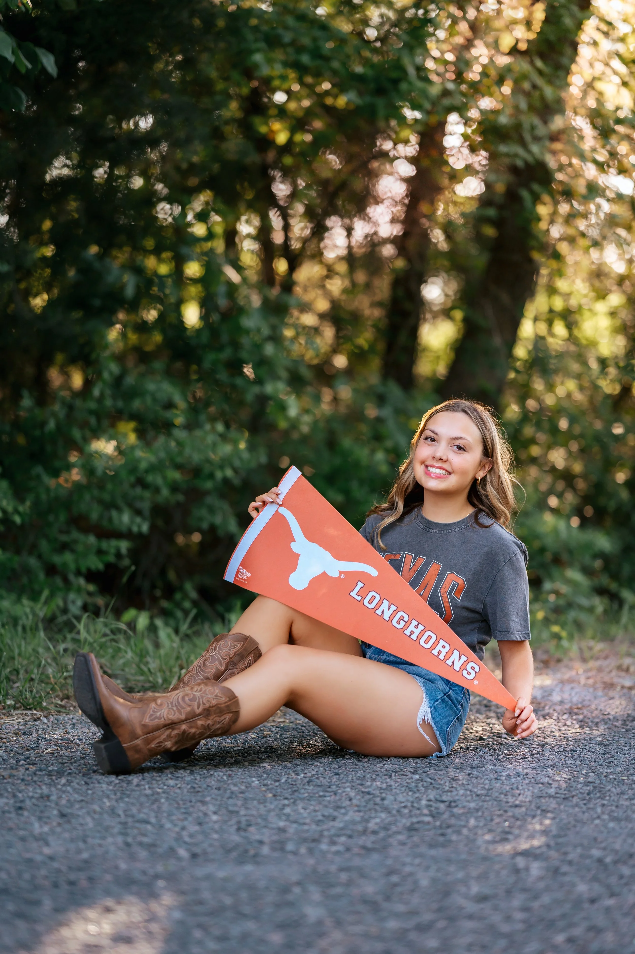 High school senior girl sits on gravel road holding a University of Texas banner, and wears denim shorts, cowgirl boots, and a gray college tee. There are trees and sunlight in the background.