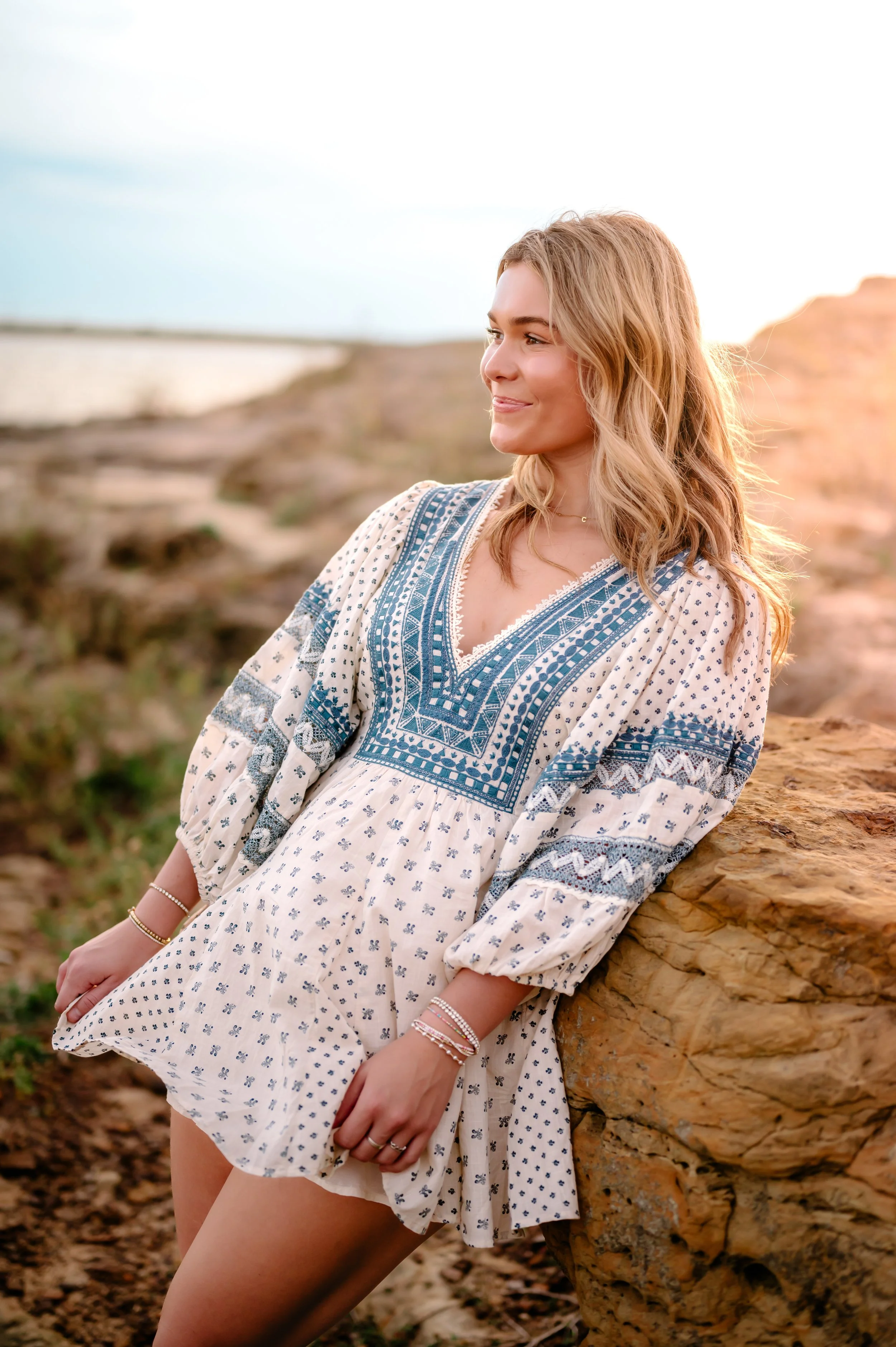 A smiling high school senior girl with long blonde hair poses against rocks by a lake shoreline. She wears a sundress, and holds the ends of it in her hands on each side.