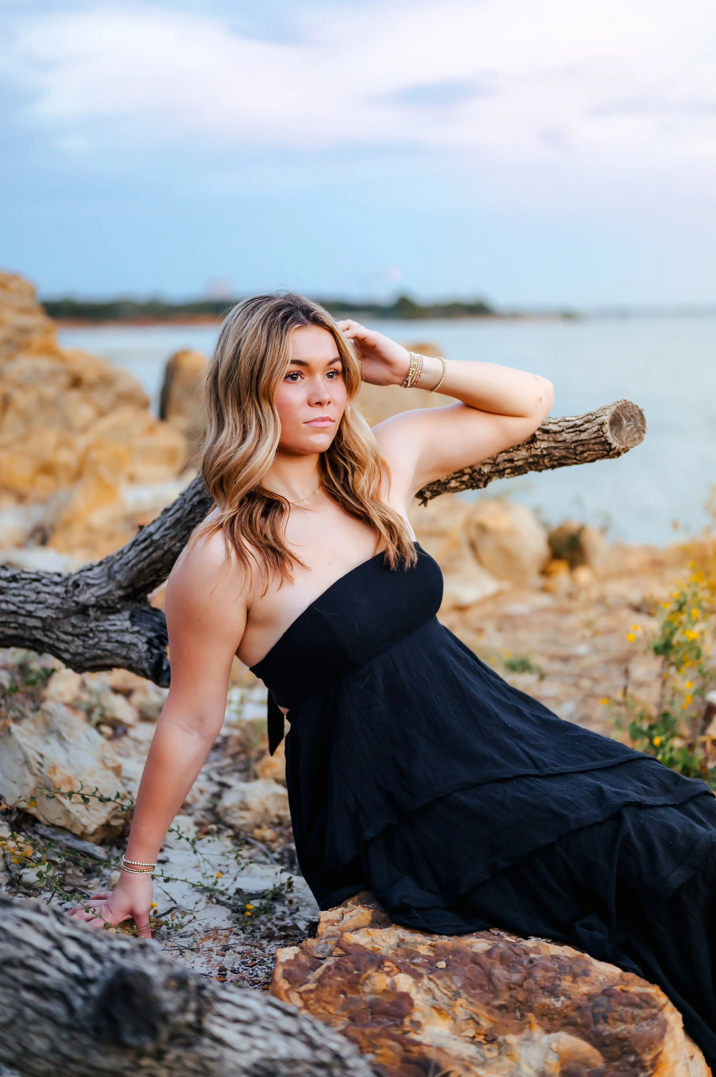 Senior girl with long hair and black dresses poses on rocky shoreline with a blue sunset sky