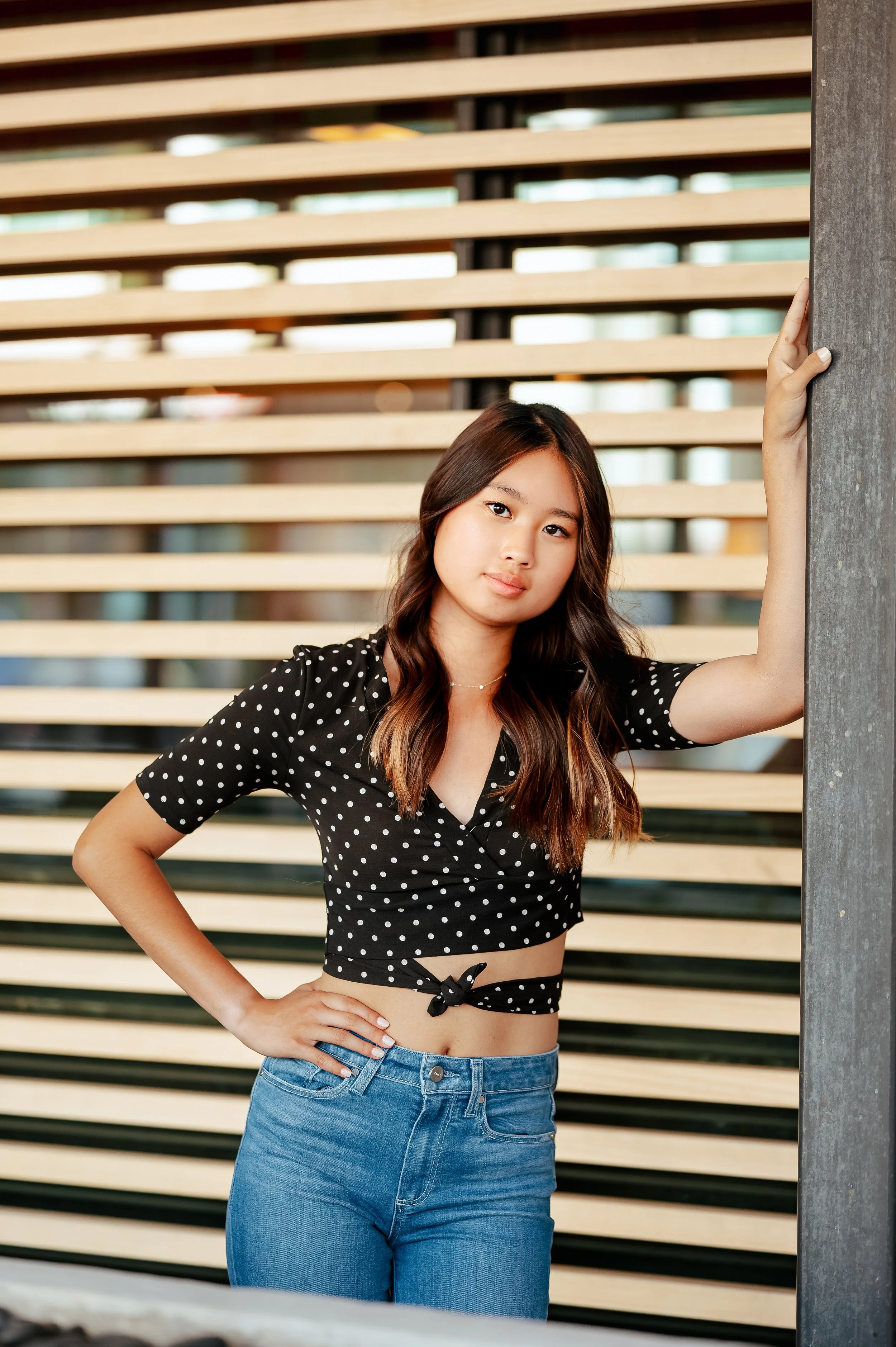 High school senior girl with long dark hair poses standing with a hand on one hip and the other against a post, with a wooden slatted background. She wears blue jeans and a black polka dotted top.