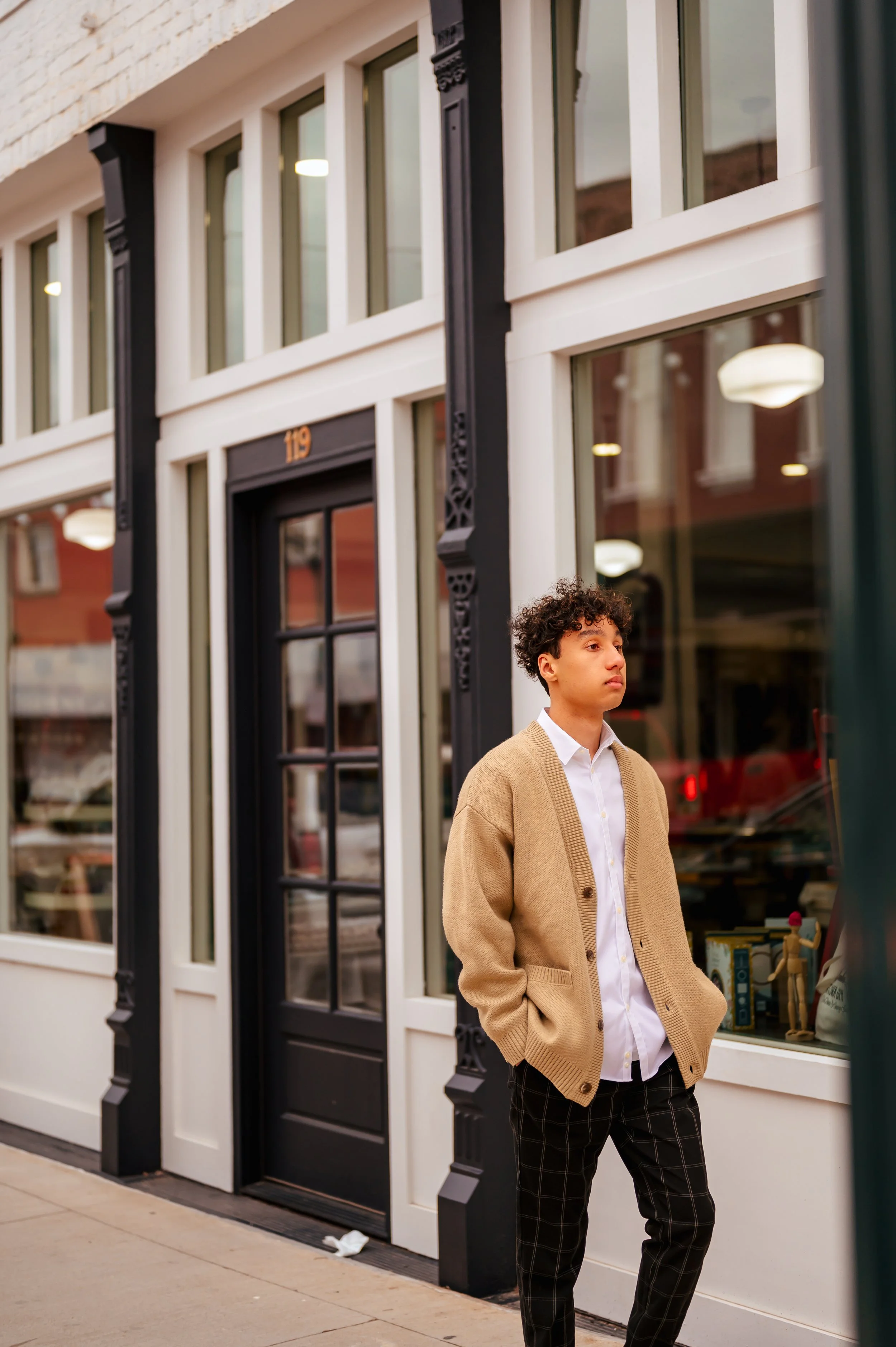 High school guy in dark pants, white shirt, and tan cardigan walks down a sidewalk with hands in pockets outside storefronts