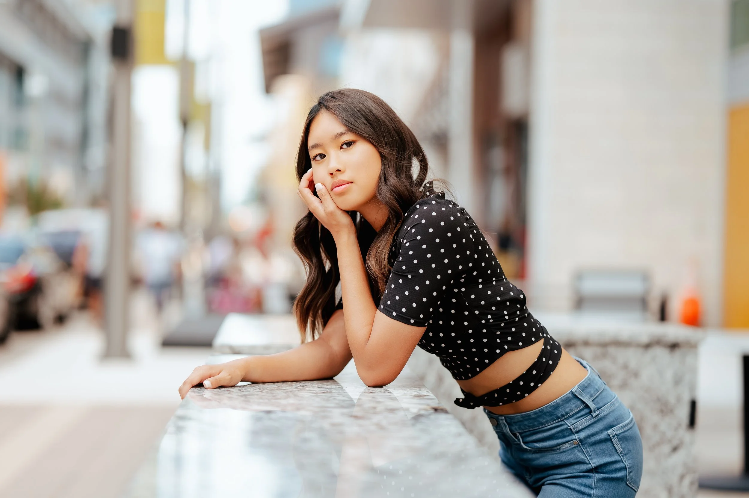 Senior girl in black polka dot top and blue jeans poses leaning forward against marble ledge in urban area.
