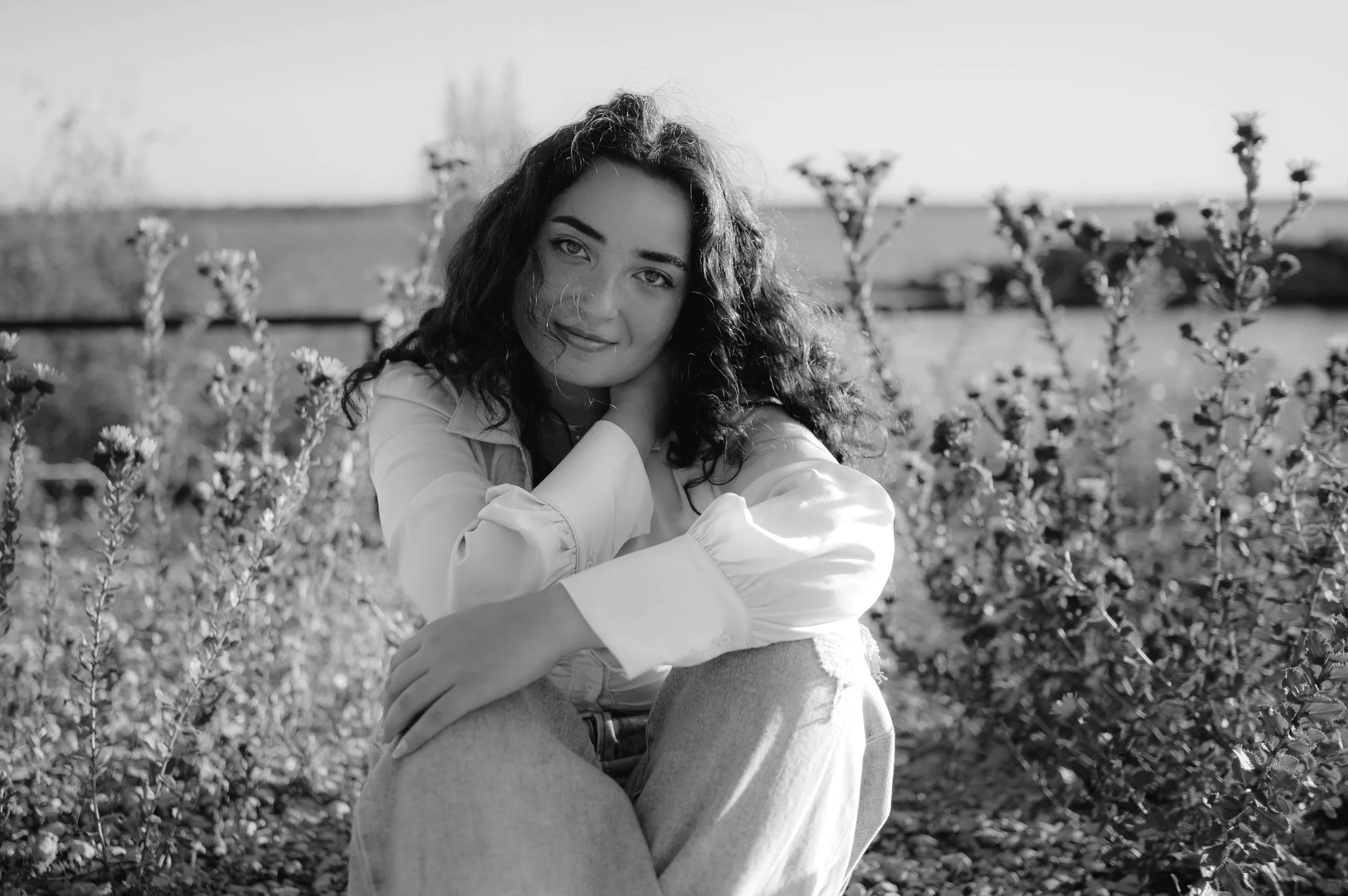 A black and white shot of a teen girl with dark curly hair sitting with her knees pulled up to her chest, amongst shrubs and flowers. She's looking at the camera with a soft smile.