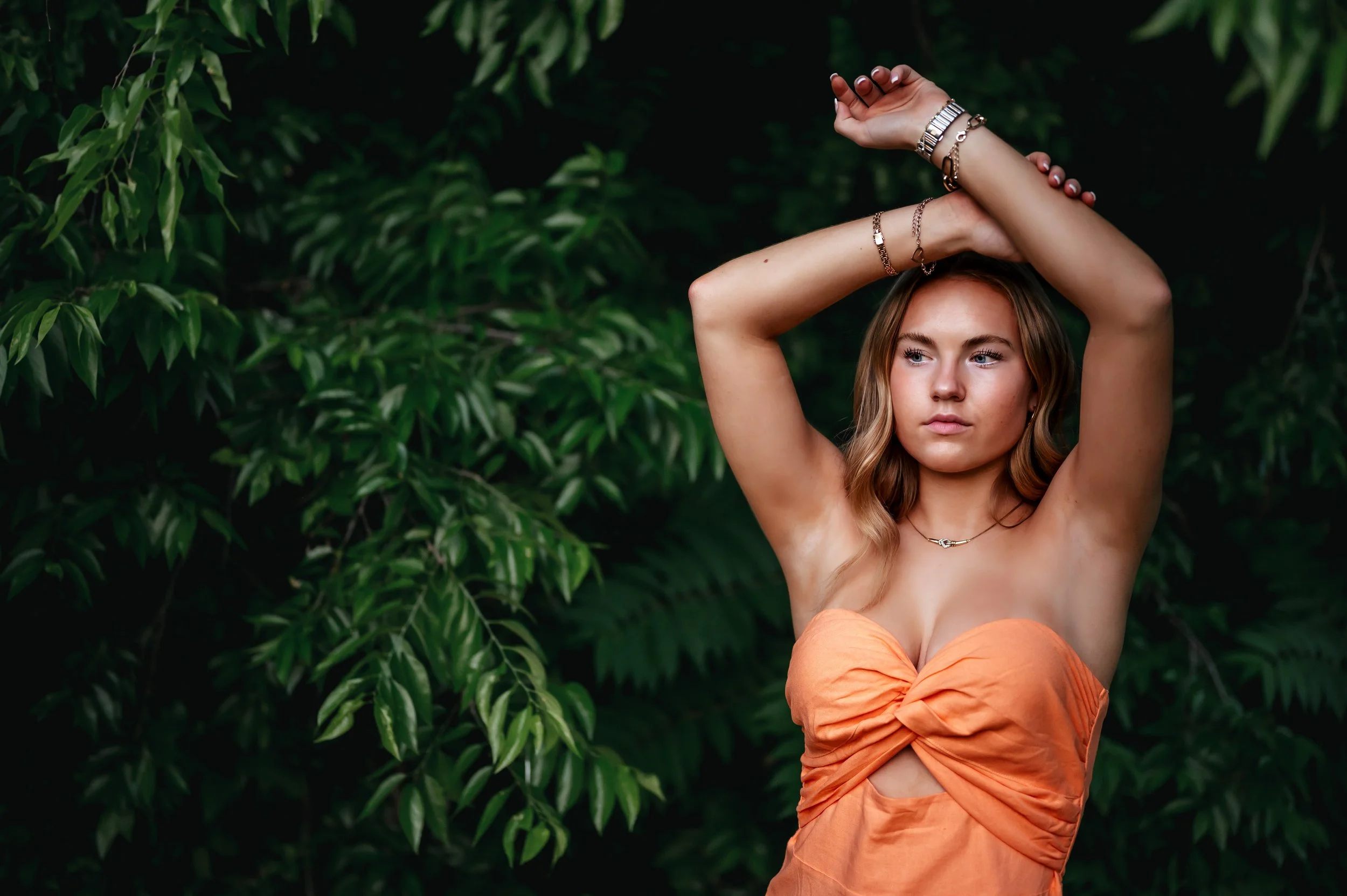 High school girl stands in front of green leafy trees with arms raised above her head, wearing an orange dress. The shot is from the waist up.