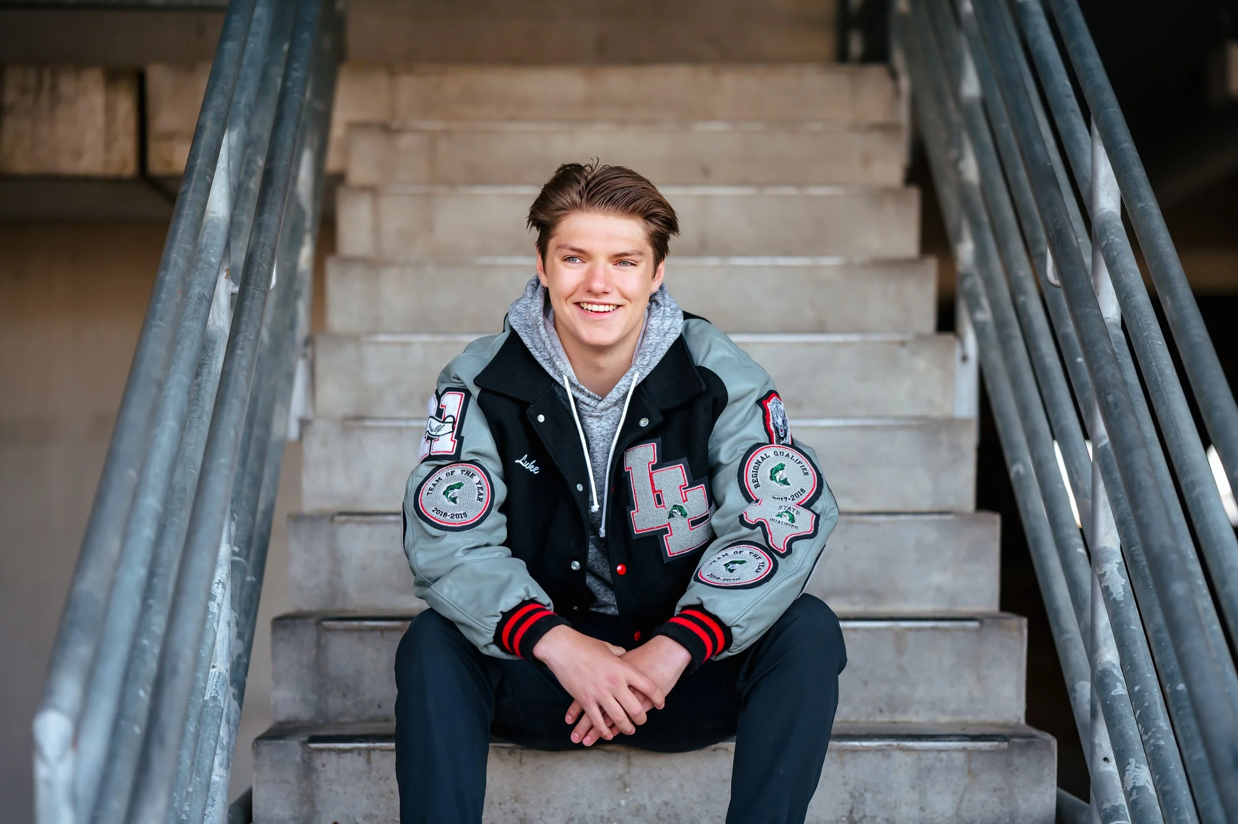 A teen boy in his letterman jacket sits on concrete stairs, smiling at the camera.