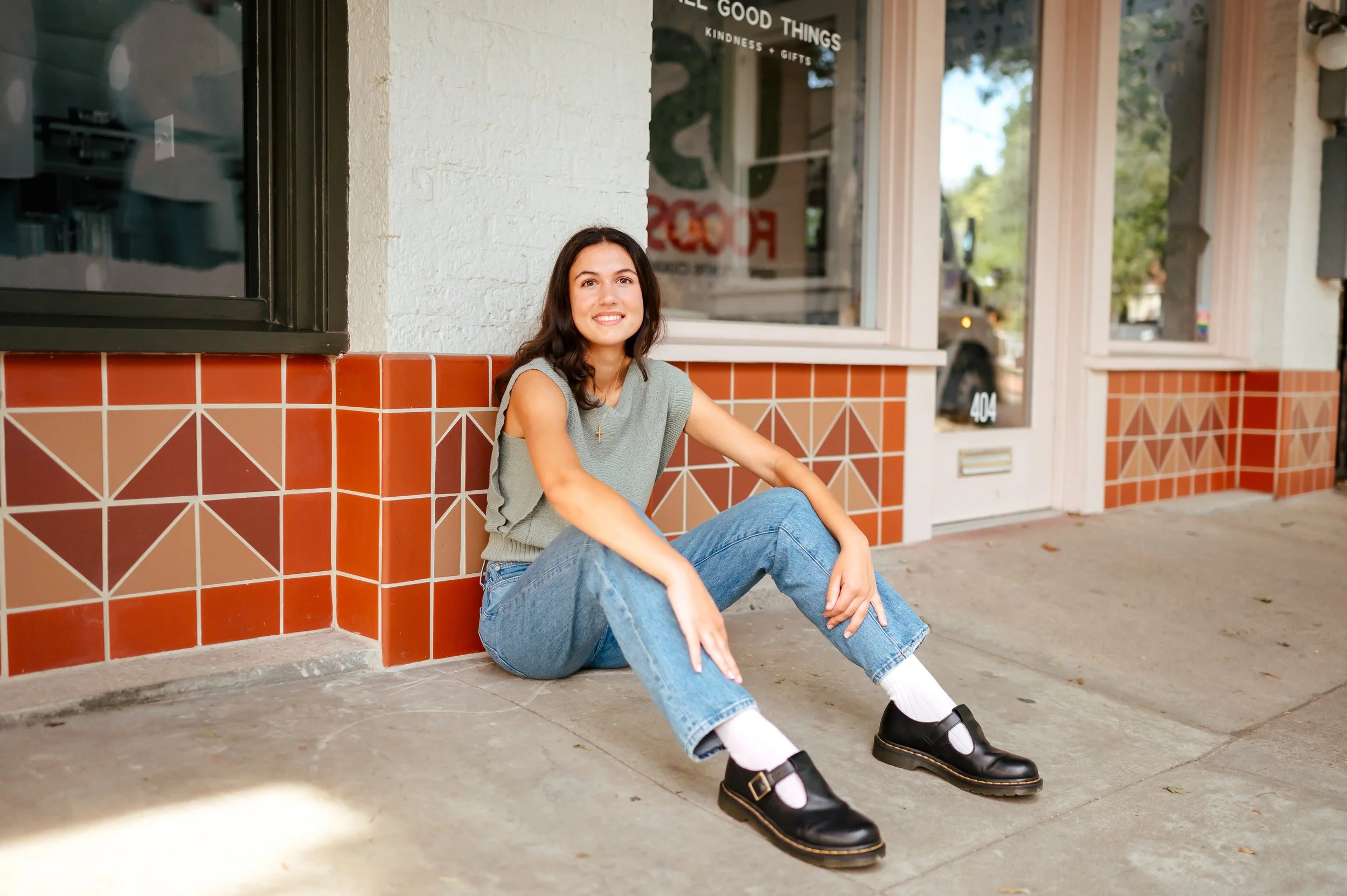 Brunette senior girl poses sitting against orange and red tiled wall, wearing jeans, black shoes, and gray sleeveless top. She's smiling, and has hands on shins, with knees up.