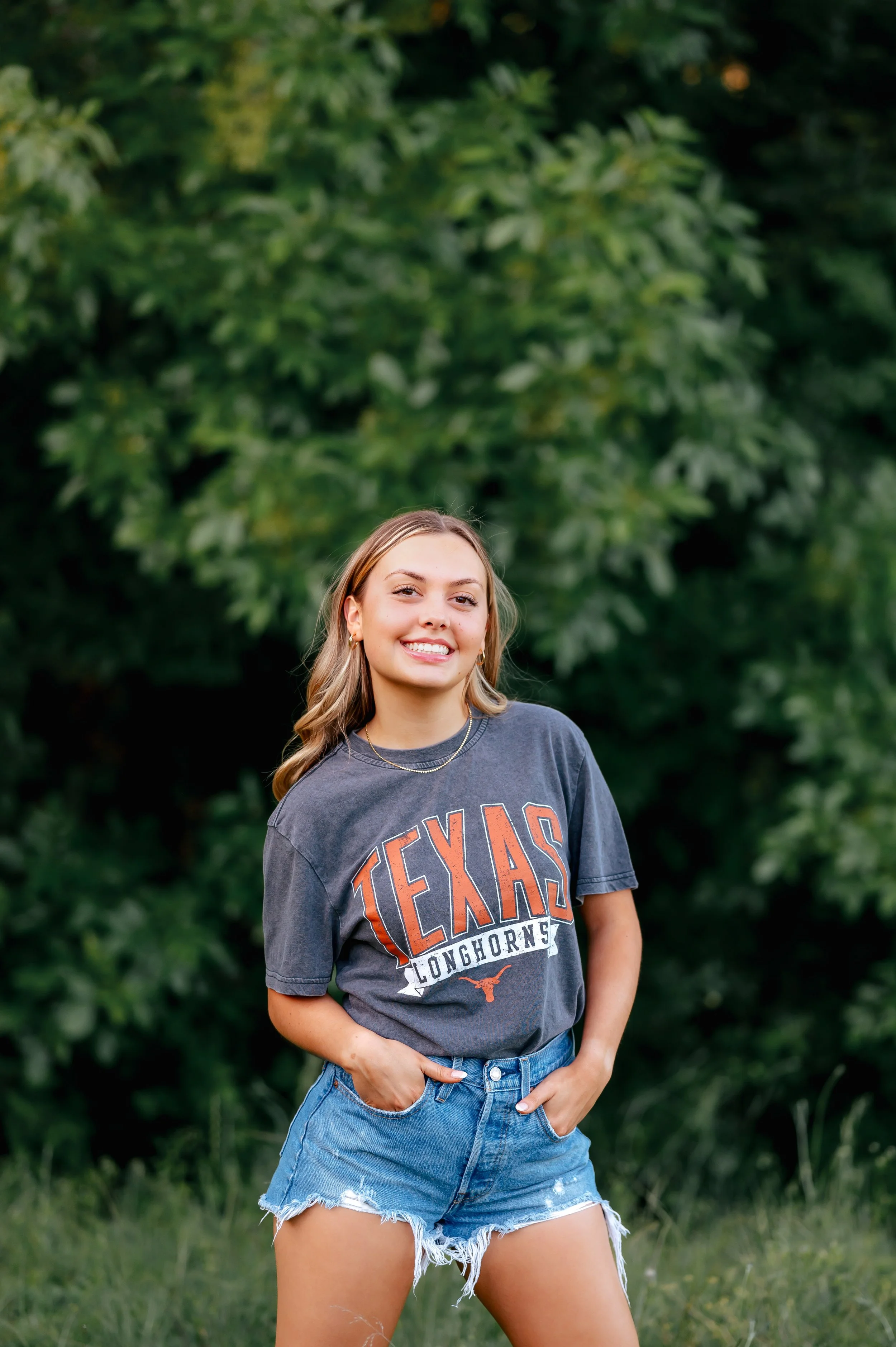 Senior girl poses in front of green trees with UT shirt on and blue jean shorts, with a smile on her face and hands in her pockets.