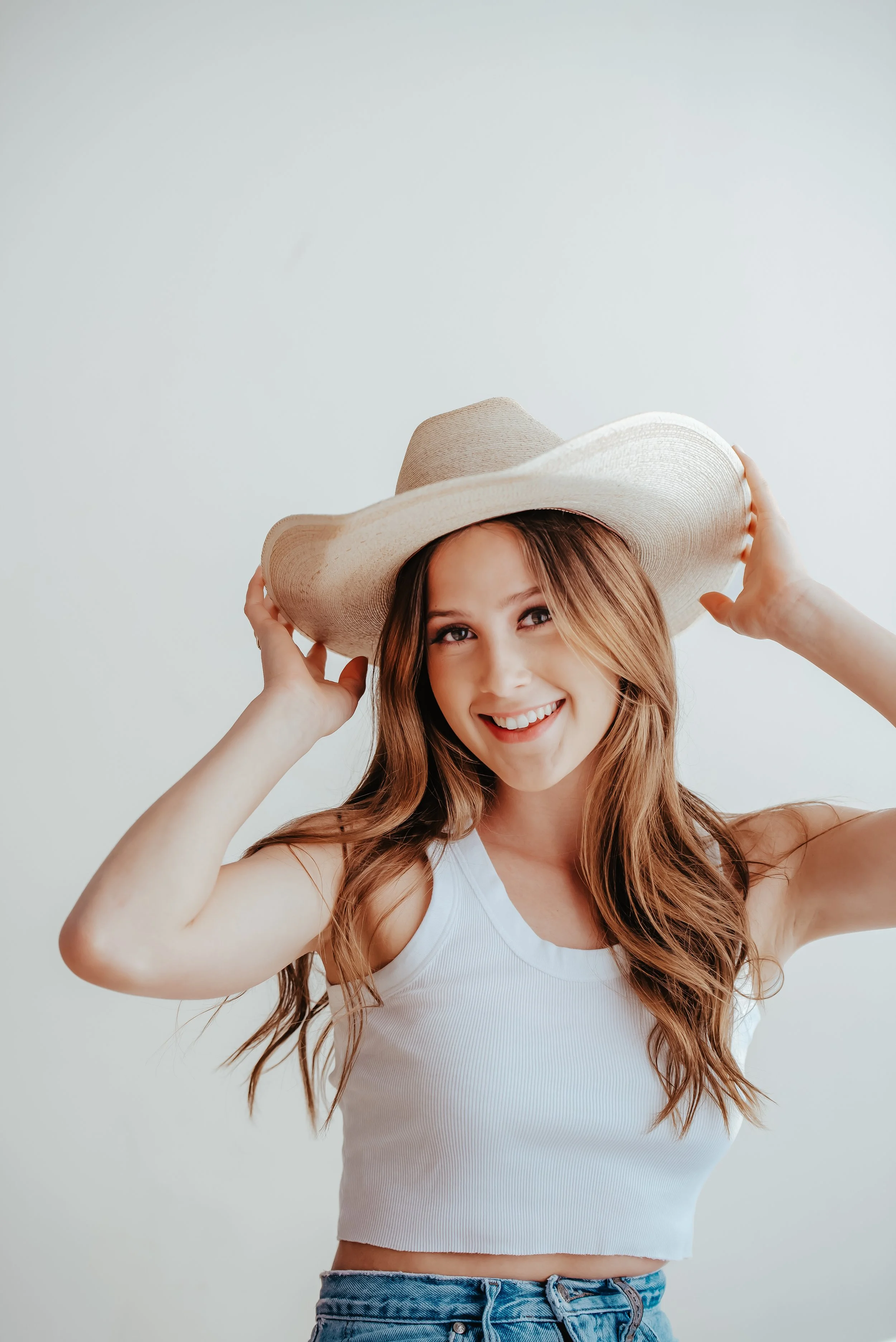 Teen girl poses with hands on her cowboy hat, with a white background in a natural light studio. She wears a white tank top and blue jeans, and it is shot from the waist up