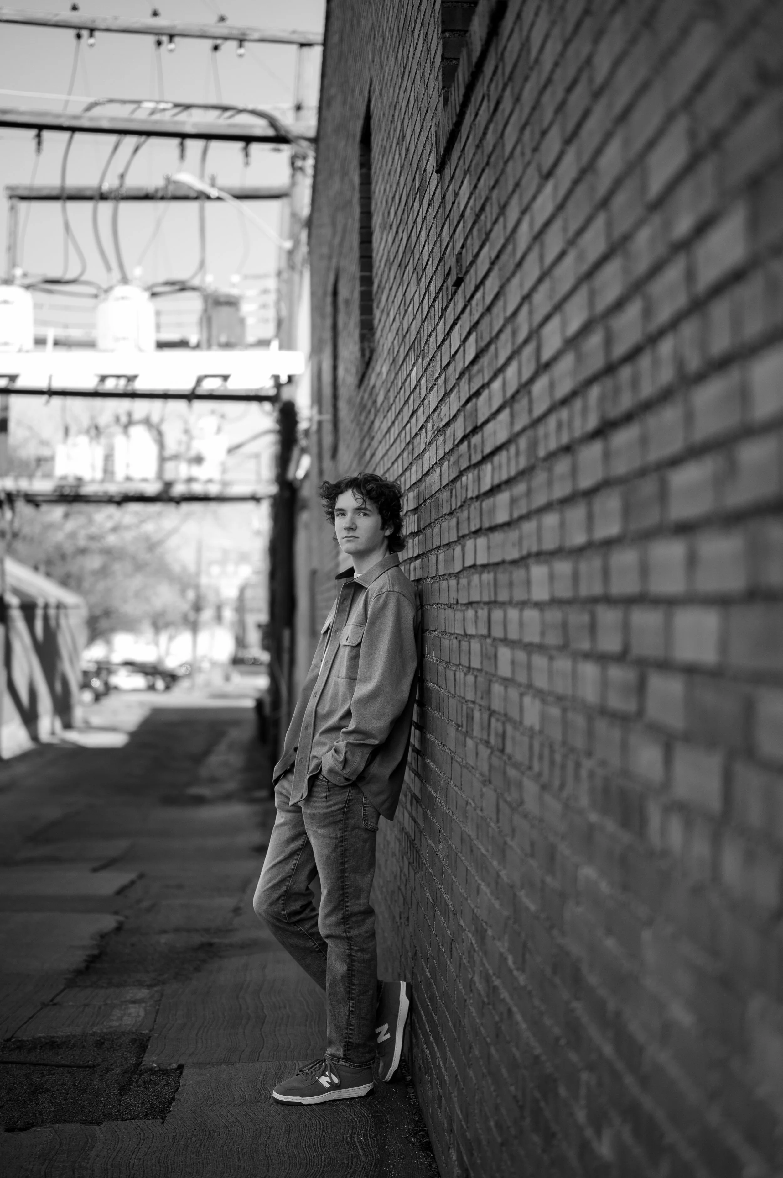 Black and white shot of senior boy leaning against a brick wall. It is a full body shot, and he's looking towards the camera, straight-faced, wearing blue jeans and a button up shirt.