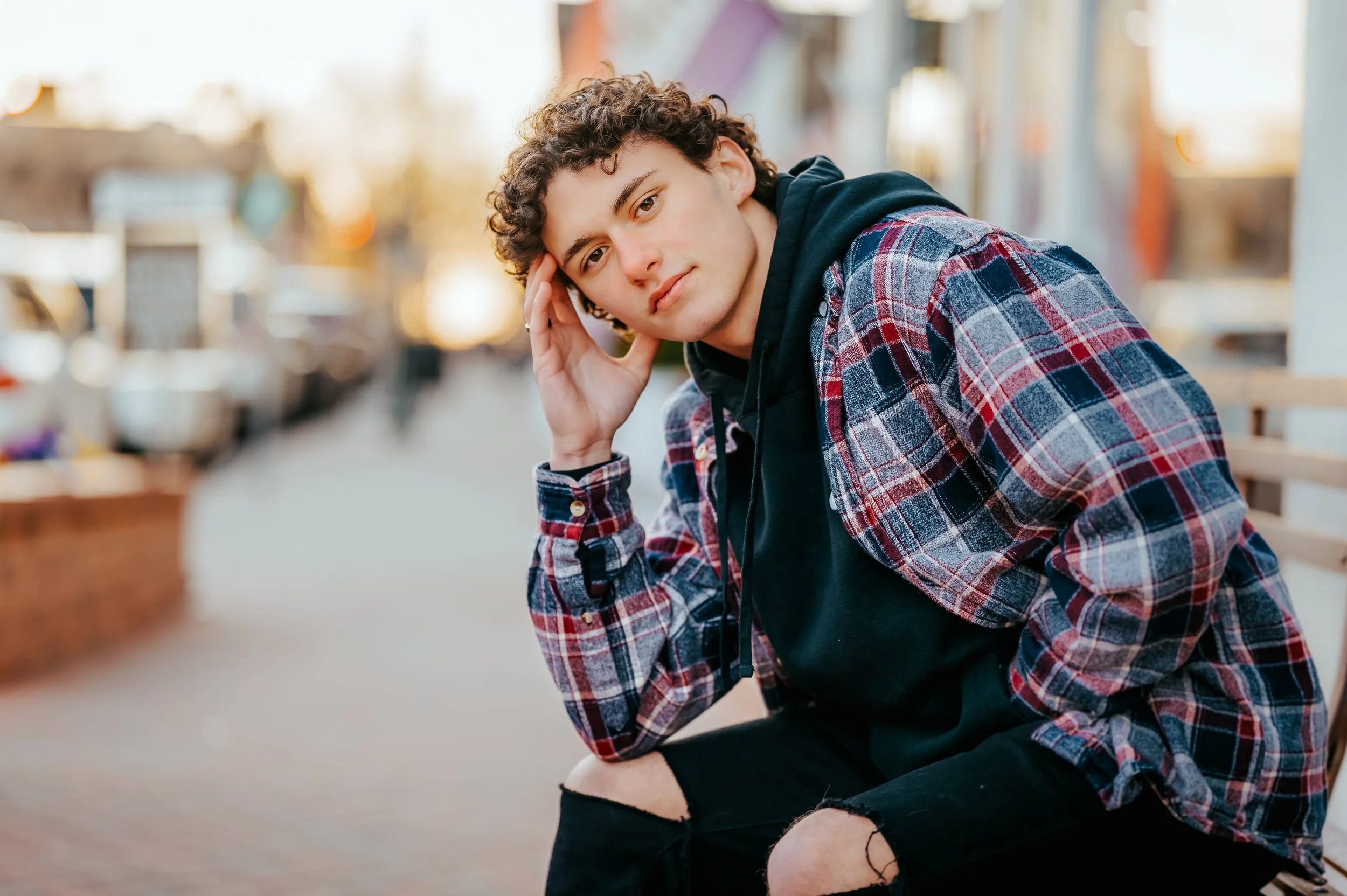 Senior boy wearing a black hoodie with plaid jacket over it and black ripped jeans sits straight-faced on a downtown bench with hand on his face
