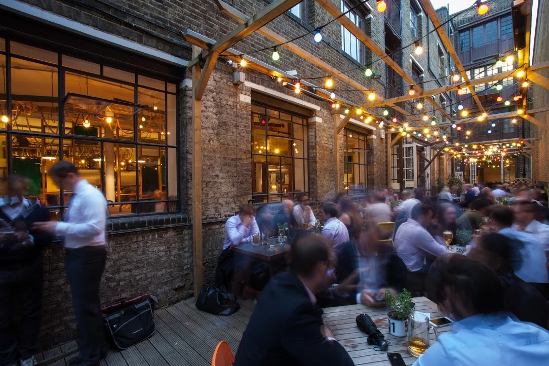 Tanner & co exterior, the yard on a busy night, lit with fairy lights hanging from a wooden framework suspended high above the tables