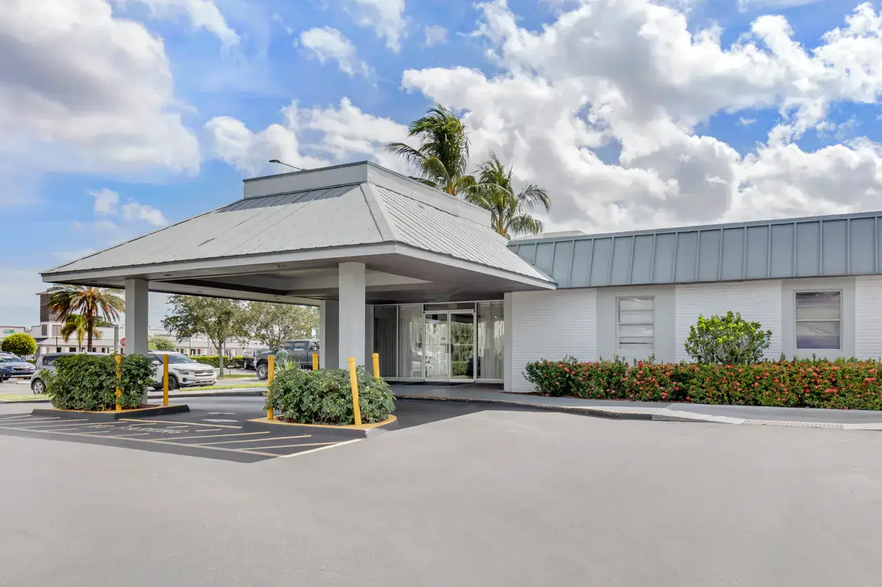 Exterior view of a building with a covered entrance and a parking lot, featuring cars and landscaping with trees and bushes, under a partly cloudy sky.