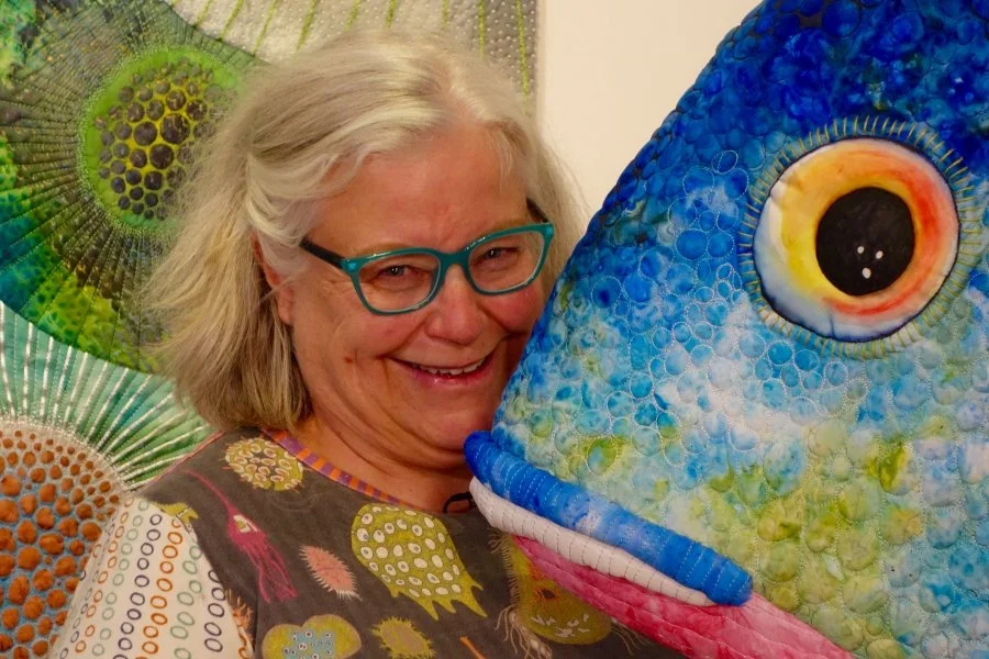A woman with gray hair, wearing glasses, smiling while hugging a large, colorful fish-shaped sculpture with detailed eyes and vivid scales.