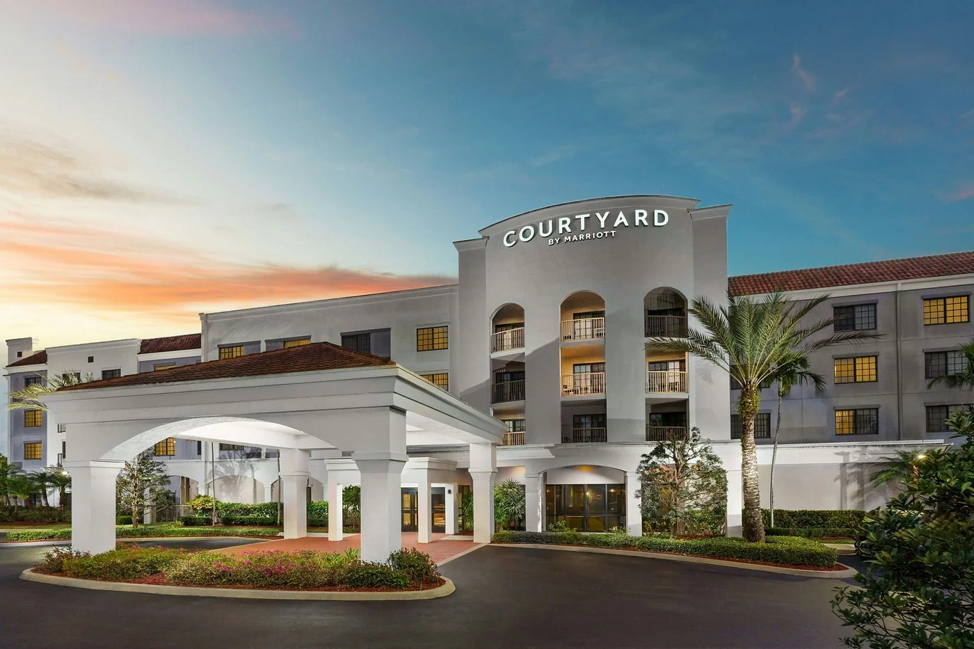 Exterior view of Courtyard by Marriott hotel at sunset, featuring a white building with palm trees and landscaped drive-up area.