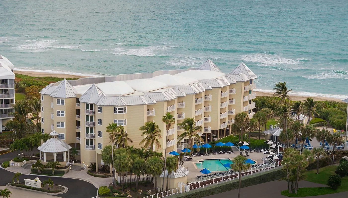 Beach resort with a large hotel building, swimming pool, palm trees, and the ocean in the background.