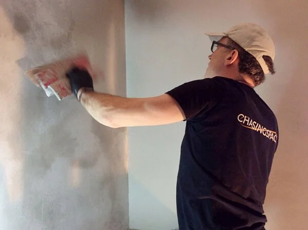 Workman trowelling a textured, grey, polished plaster wall with a trowel, viewed from the side.