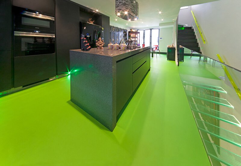 A contemporary kitchen interior featuring a bold, vibrant lime green resin floor, perfectly offset by dark grey units and a glass staircase.